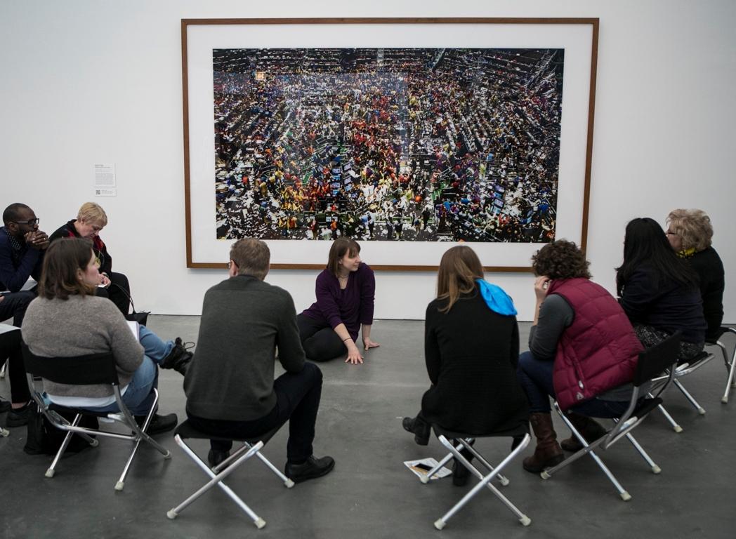 A guide sitting in front of a large photograph talks to a small group of seated adults.