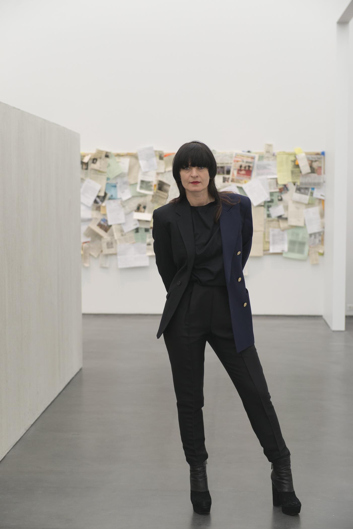 Portrait of a woman with long brown hair, blunt bangs, red lipstick and dressed in black standing in an exhibition at the MCA.