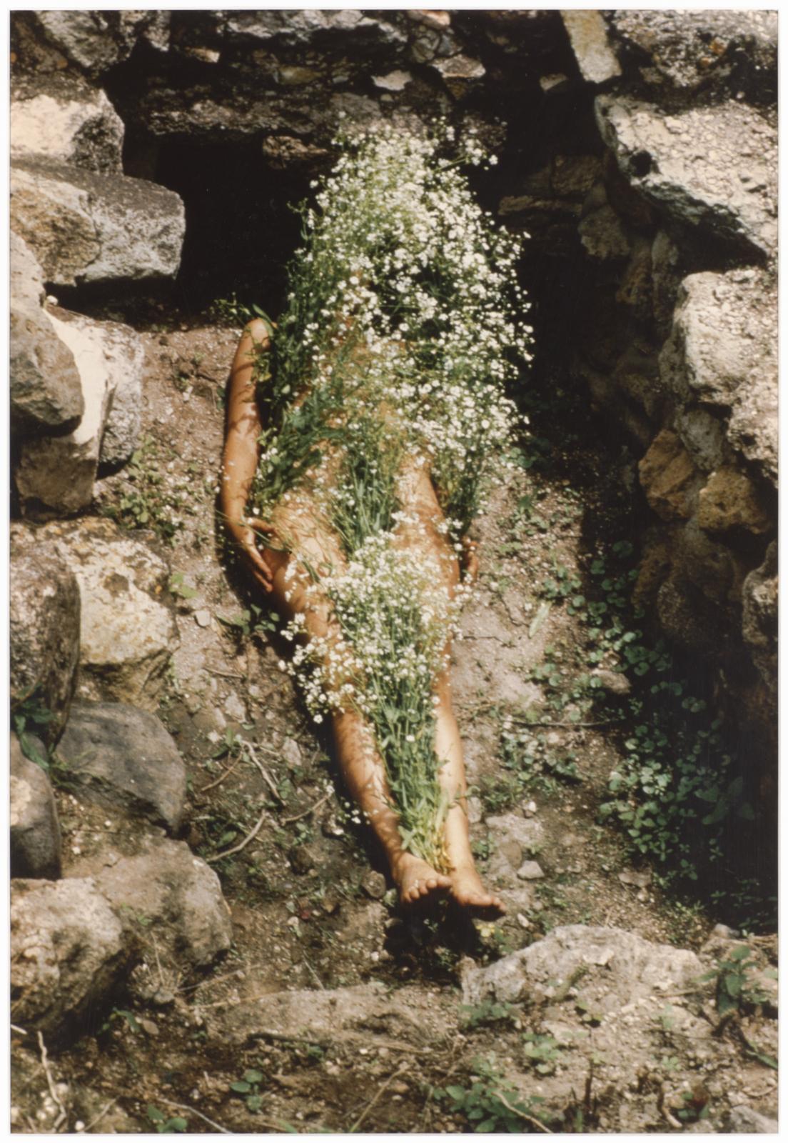 A person lays down in a stone-lined grave with flowers covering their body.