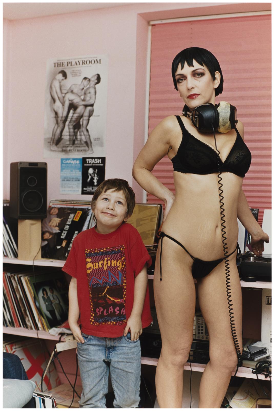 An adult wearing large headphones and black lingerie stands confidently next to a smiling child in a t-shirt and jeans. The pink wall behind them features a shelf of records and audio equipment, and a poster of nude male figures.