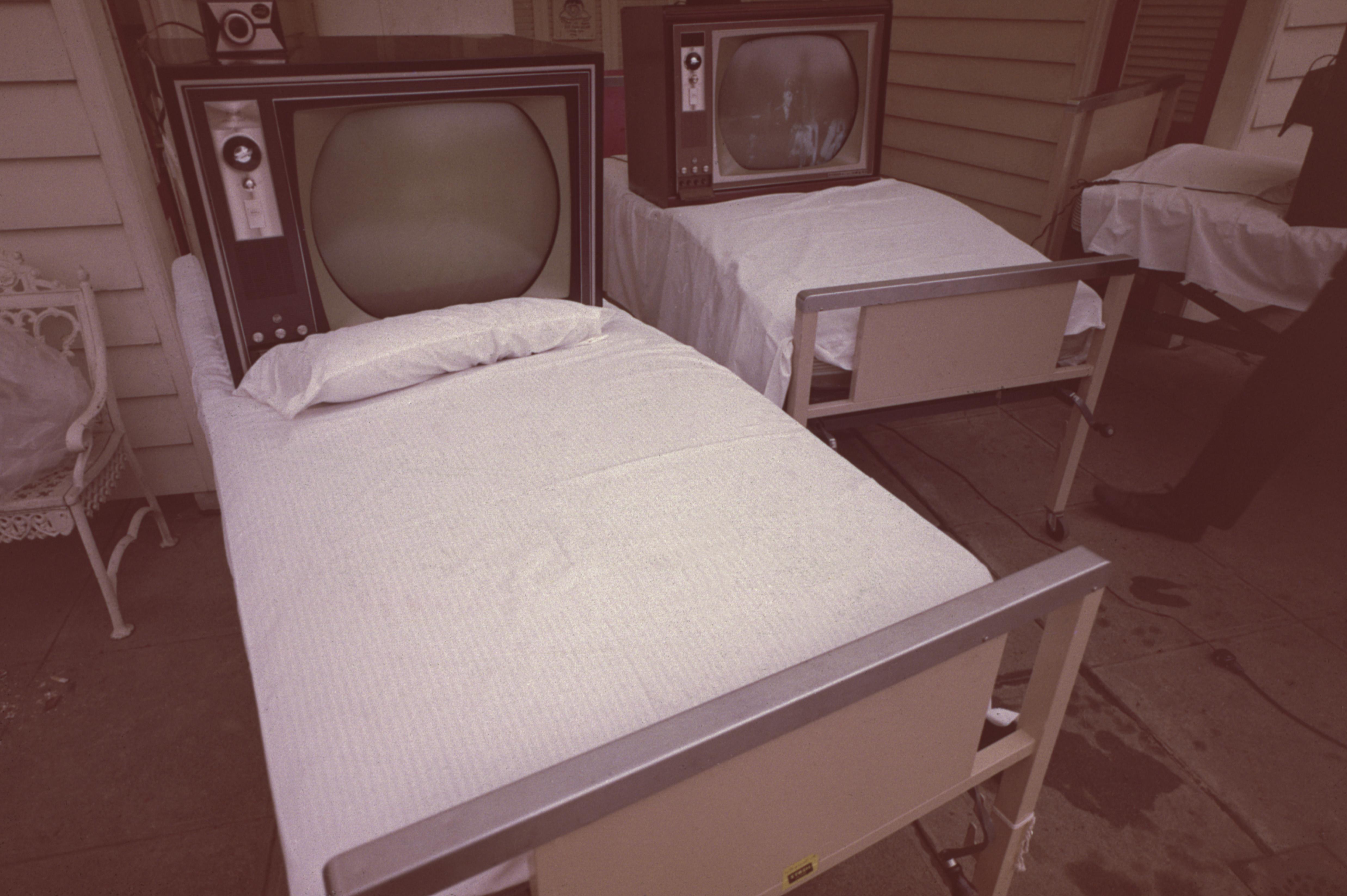 Two old hospital beds made up with white sheets are installed with 50s-style TV sets on the pillow of each bed.