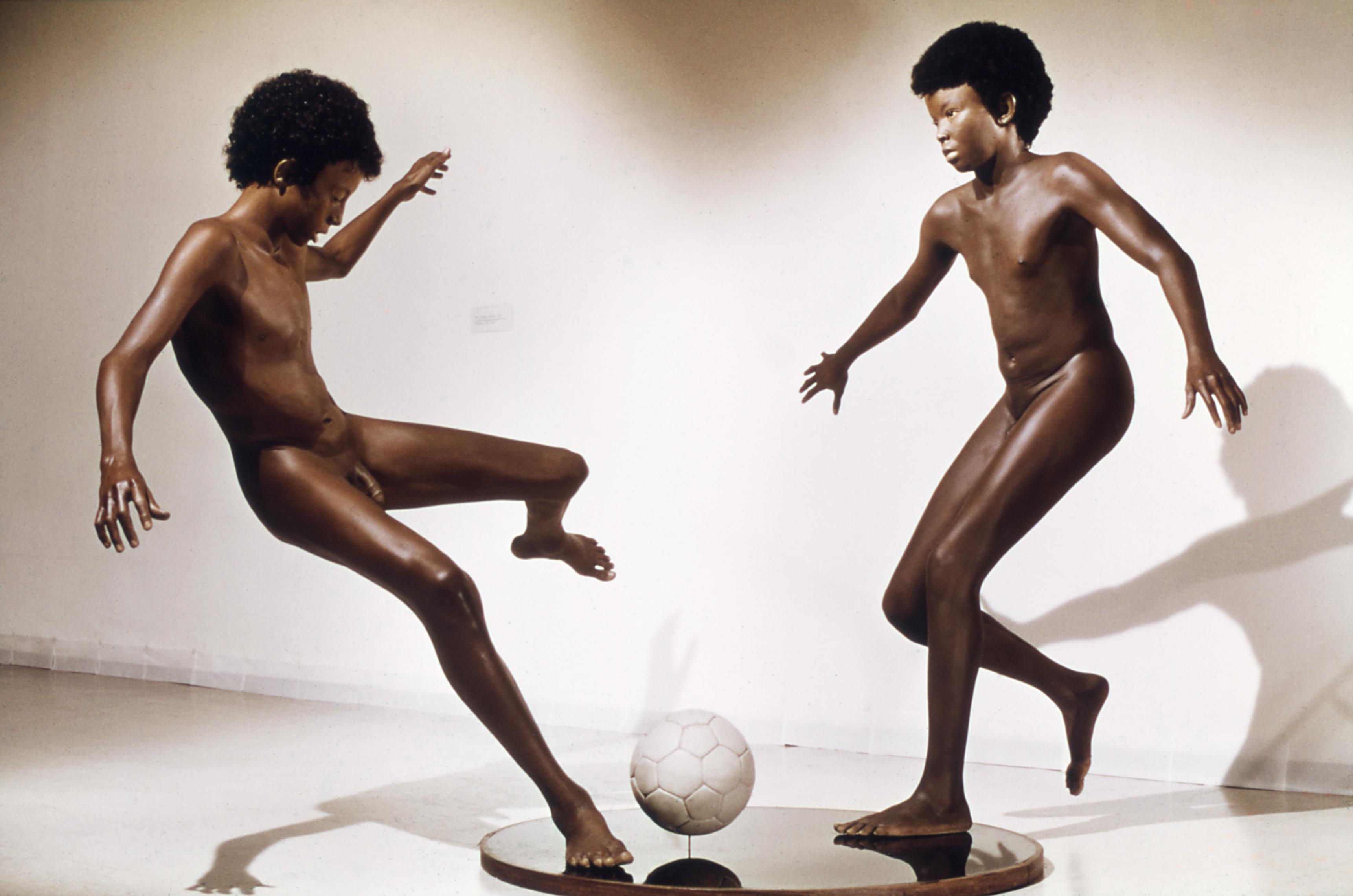 Life-size sculpture of two young nude boys with afros play soccer. One is running up to the ball, the other winds up for a kick.