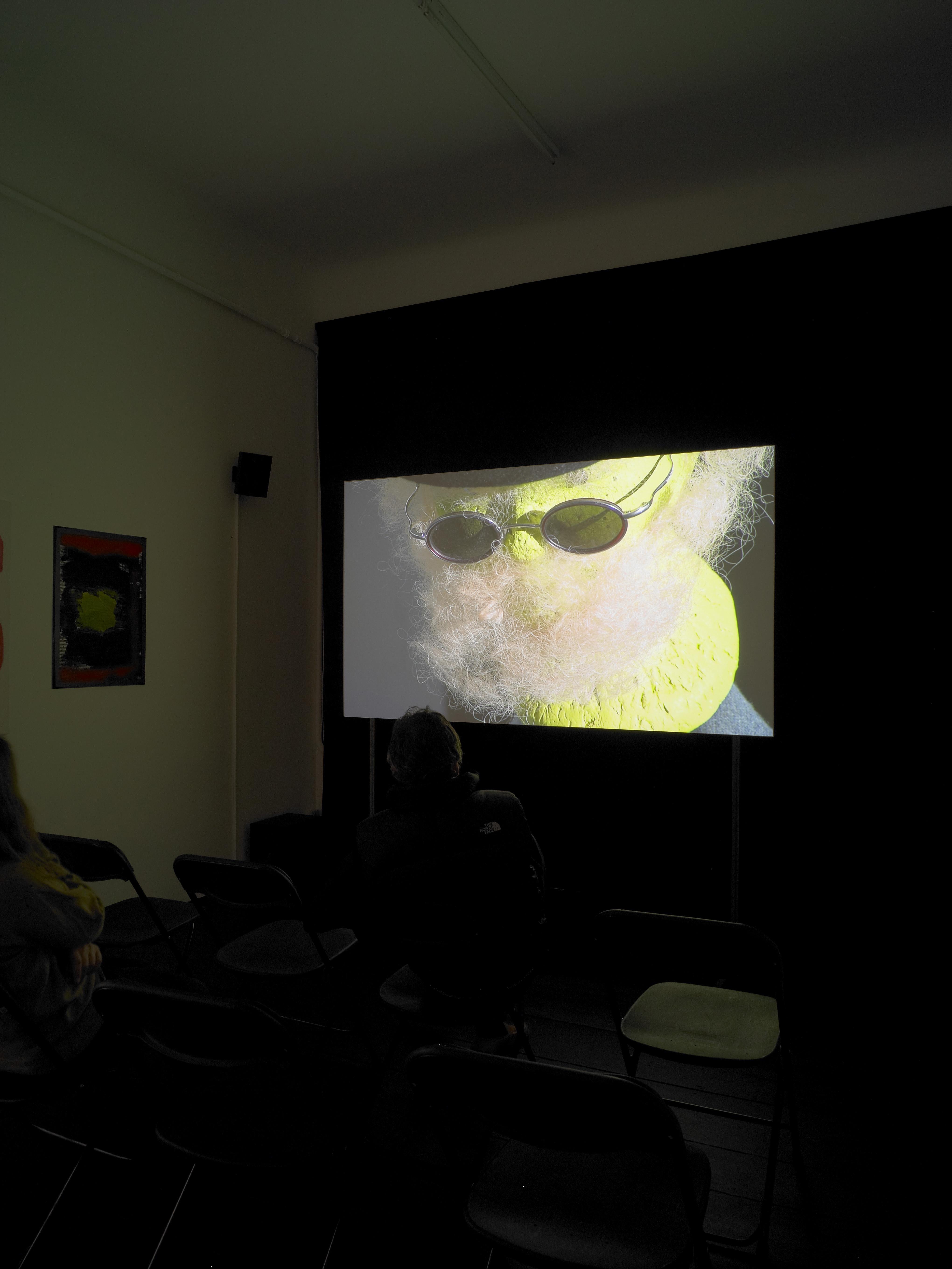 Installation view of video projection: a bright yellow object with sunglasses and tan fuzz that resembles a beard