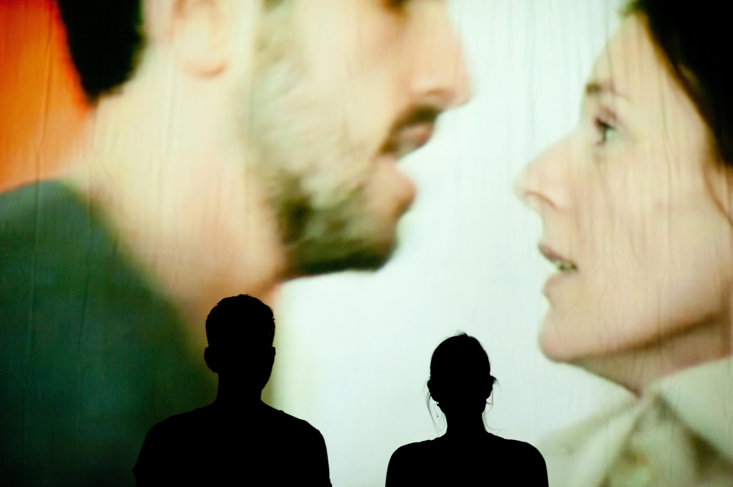 Silhouettes of two viewers watching a large video installation of a man and woman mid conversation.