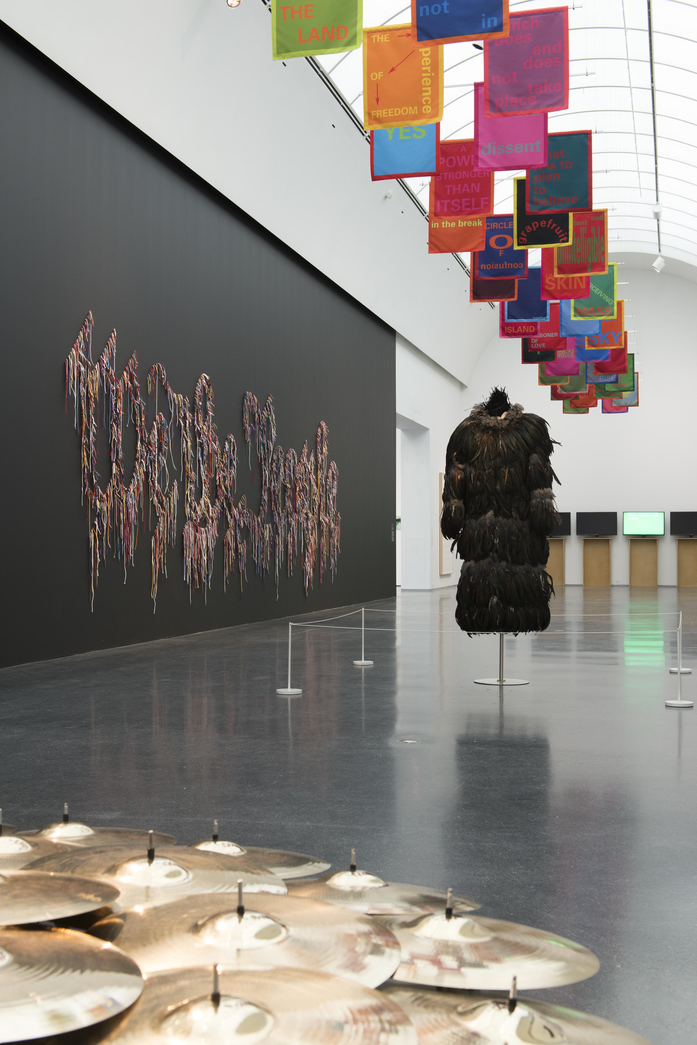 A feathered coat is displayed in the middle of a gallery, cymbals are in the foreground, and "We the People" is written on the black wall. To the left, colorful banners hang from the ceiling.