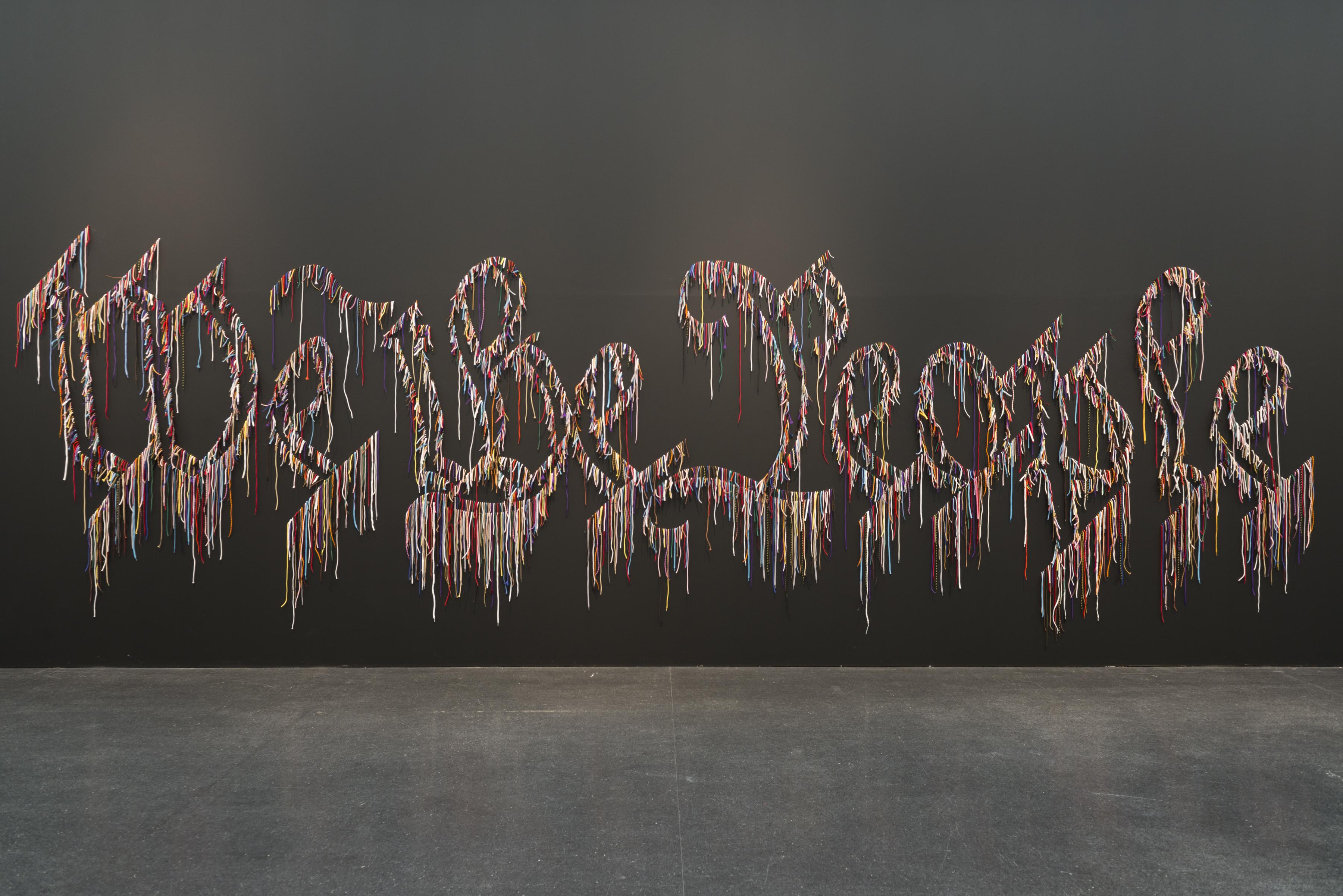 A multi-colored outline of Gothic-style letters spelling"We the People" is installed on a solid black wall. The letters appear to be dripping.