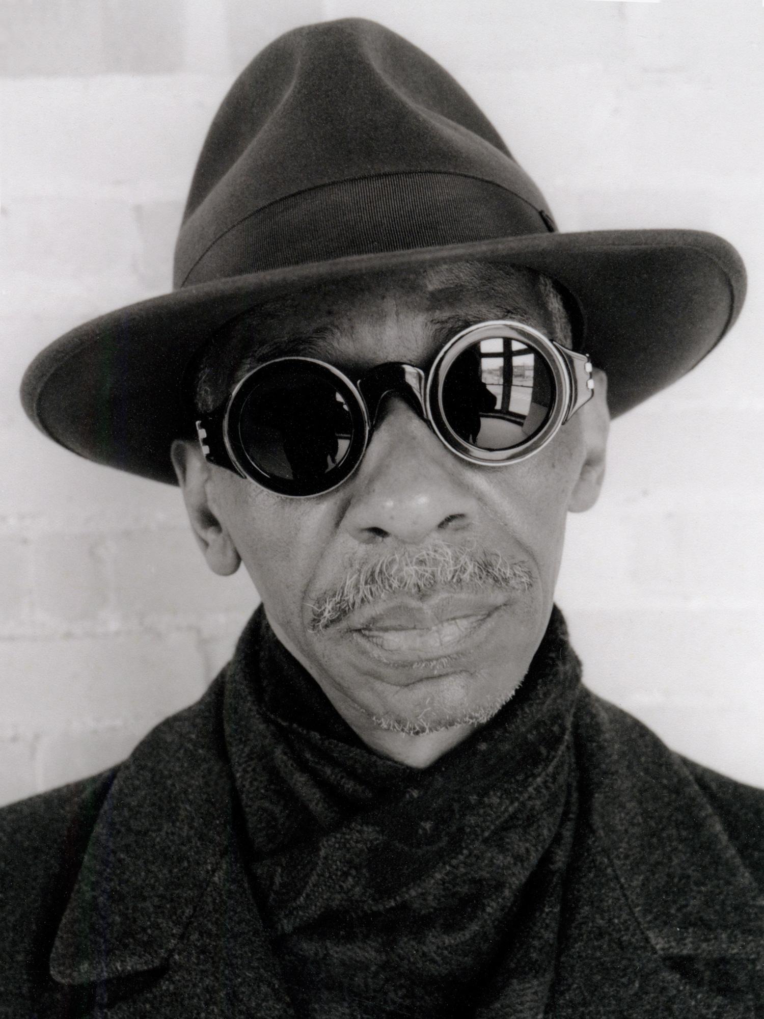 A black-and-white portrait shows an older man wearing a trilby hat and distinctive round sunglasses.