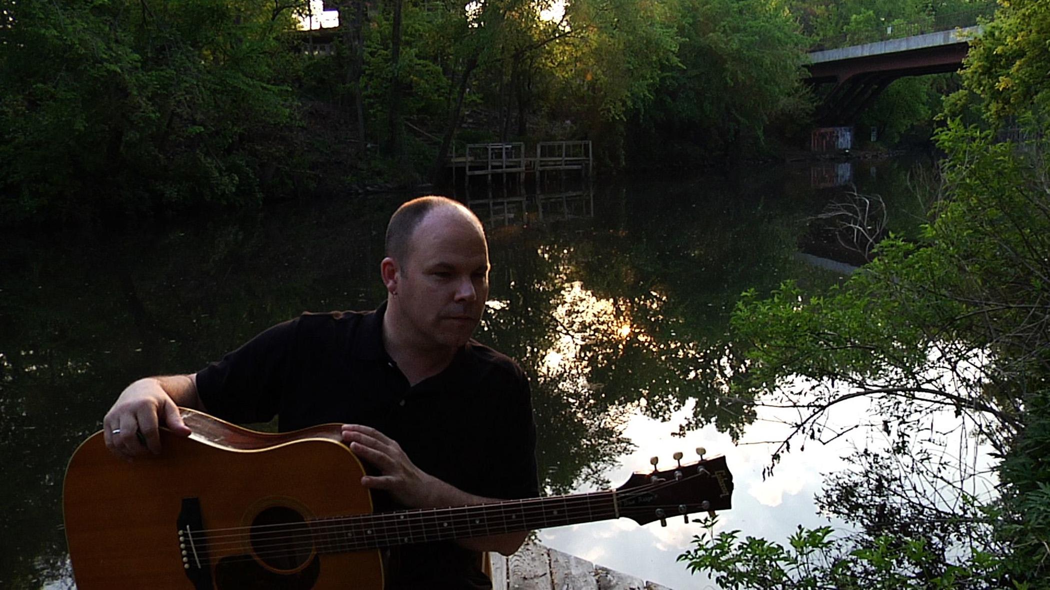 A man holding a guitar in front of a secluded tree-lined body of water