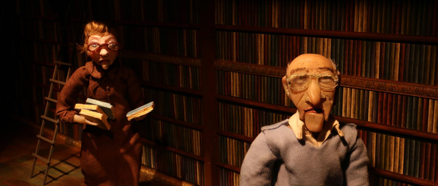 Puppets of an old man and woman walk through a dark room past a wall lined with books.