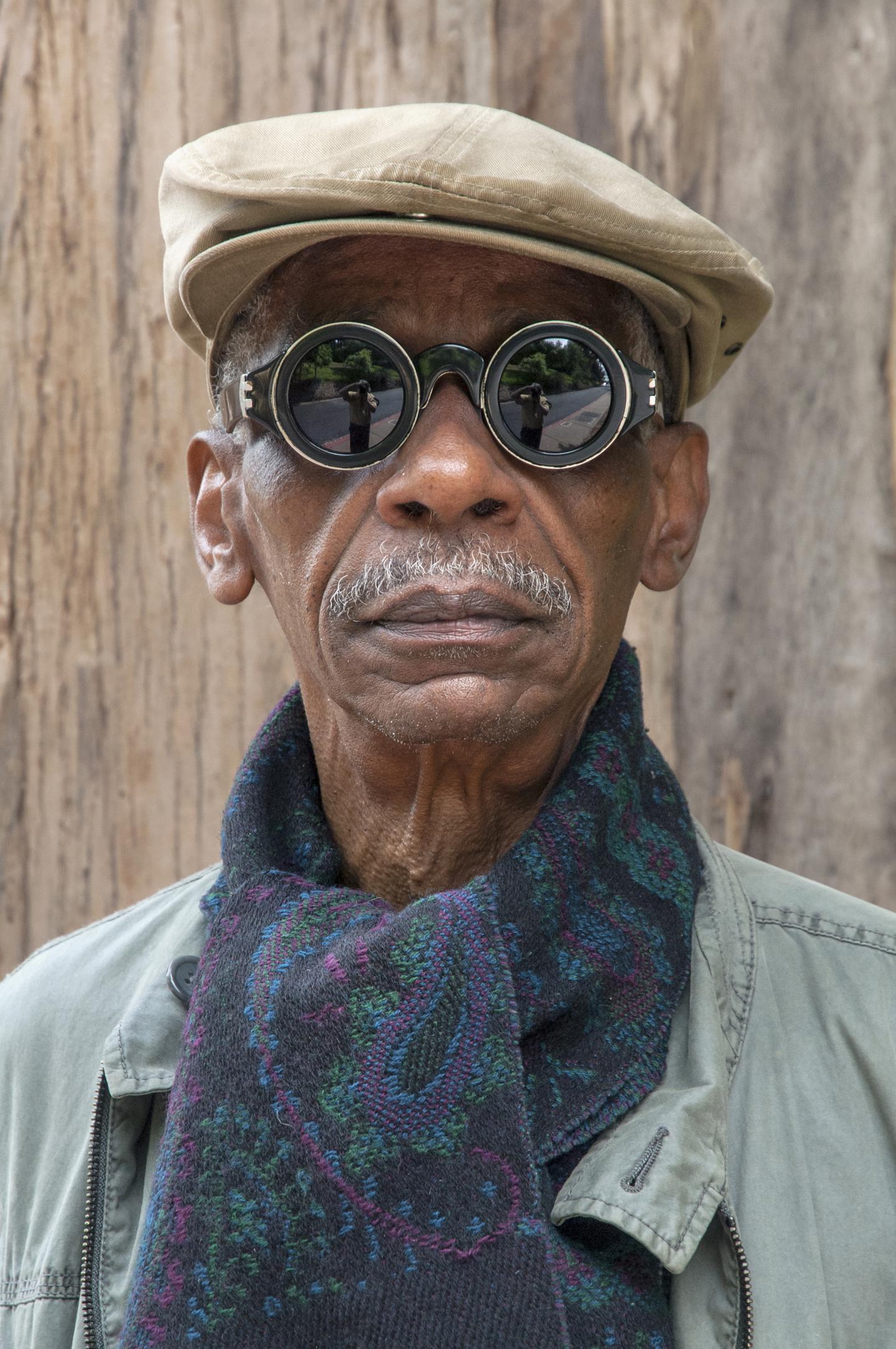 A photographic portrait shows a dark-skinned older man wearing circular mirrored sunglasses, a khaki flat cap, and a multicolored scarf.