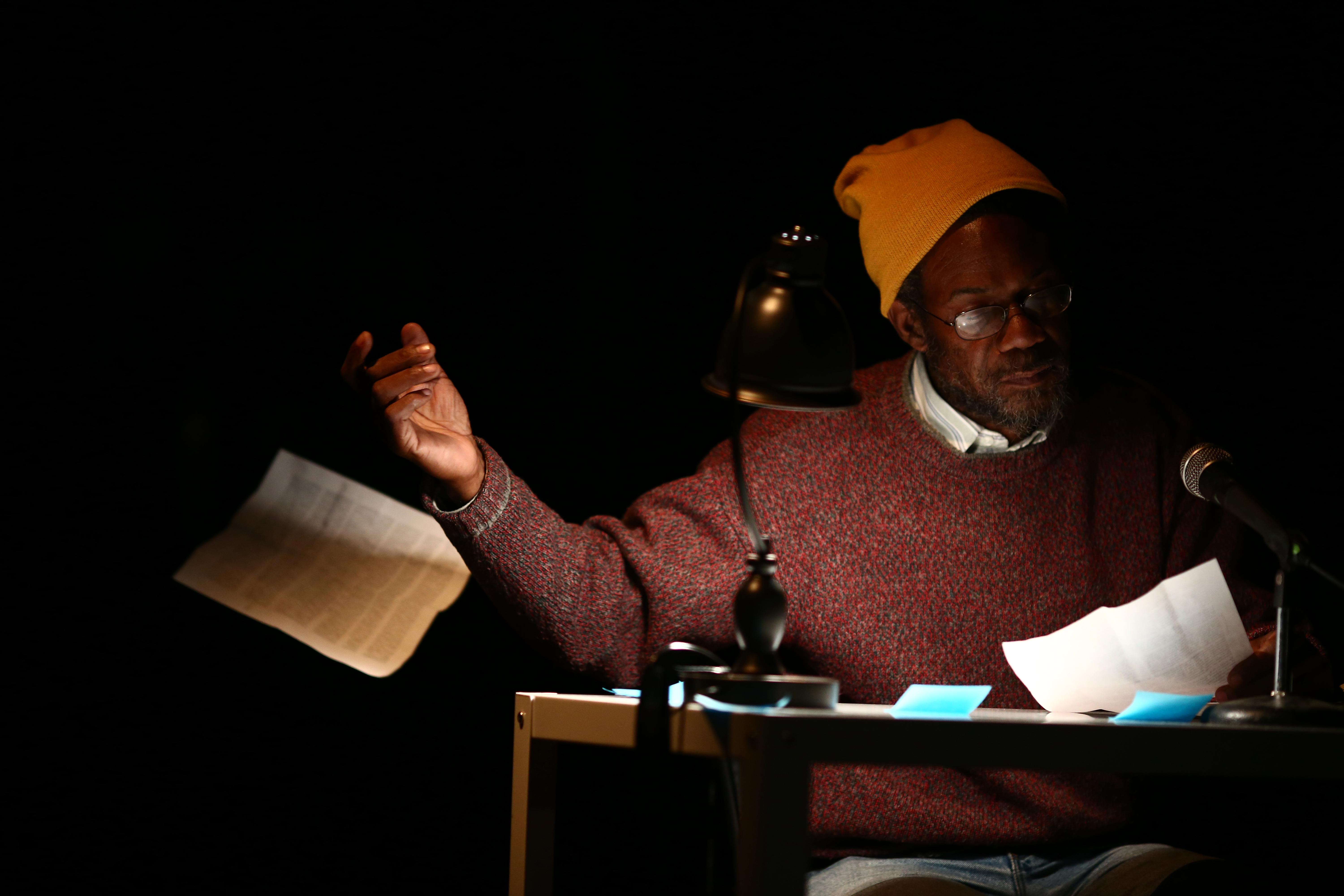 A dark-skinned older man sits at a table illuminated by a desk lamp on an otherwise pitch-black stage. He looks at one sheet of paper and tosses another to the floor.