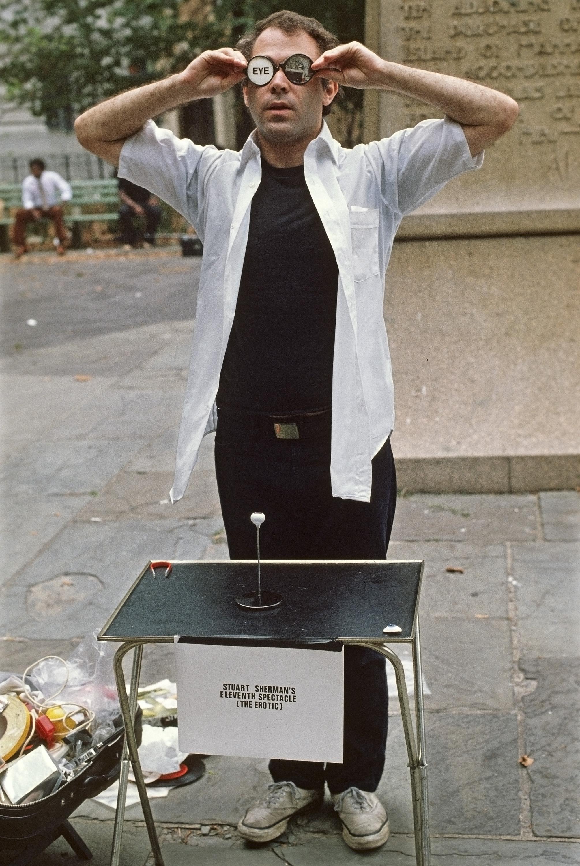 A performance still shows a man in a public park putting on round glasses that contain pieces of paper in place of lenses.