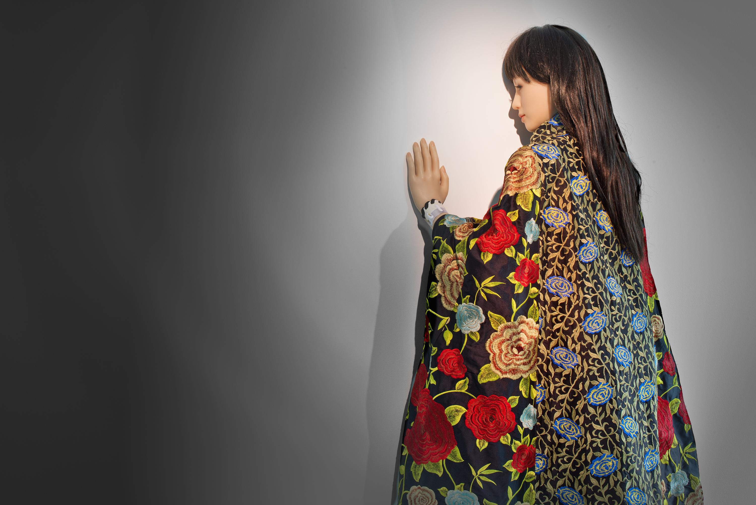 A female doll with long dark hair wearing a floral kimono stands with her back to the viewer, her palm pressed against a white wall.