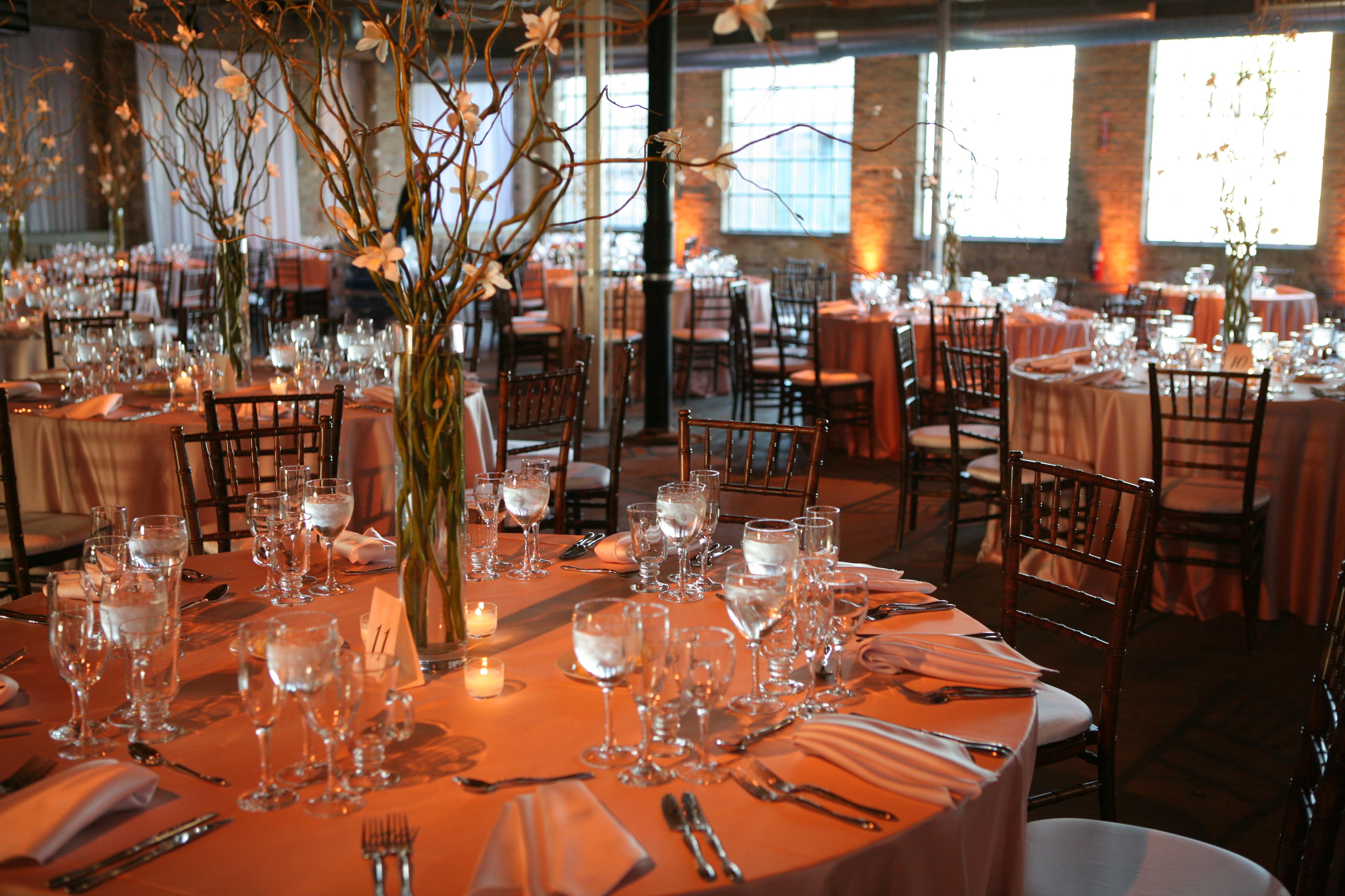 A brick-walled room with large warehouse windows is set for an event with many linen-covered roundtables. Each table is set for dinner lit with candles creating a warm glow and features an elaborate centerpiece of tall twisting vines and white flowers.