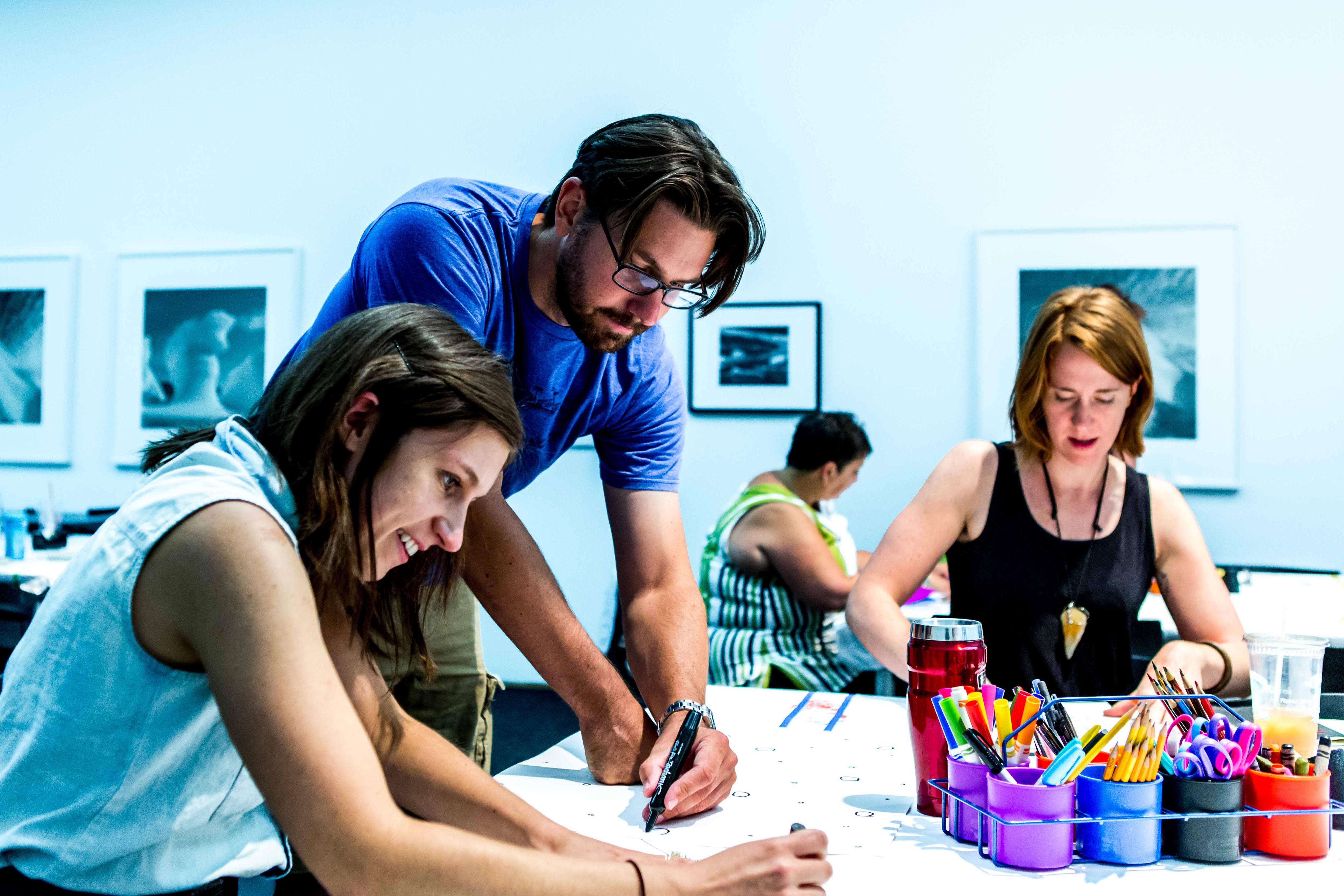 Three adults drawing on a table with framed artworks in the background.