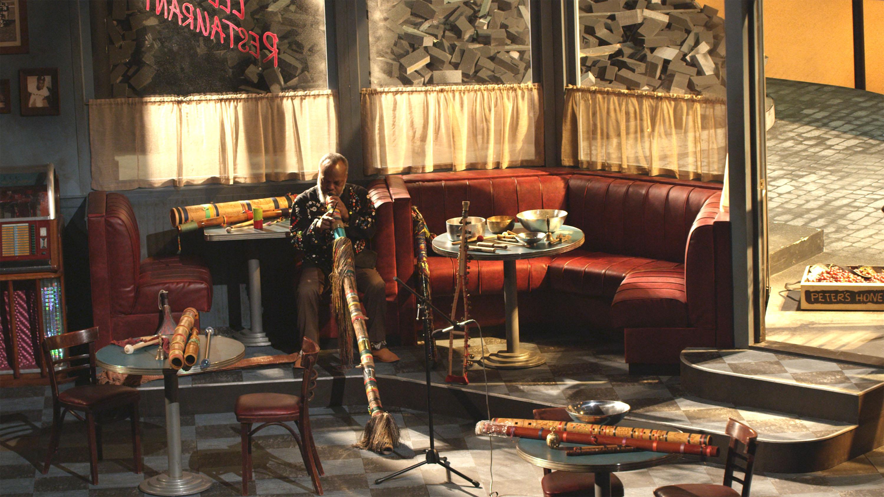 A video still shows a man playing a didgeridoo in a cafe with multiple wind and percussive instruments on tabletops.