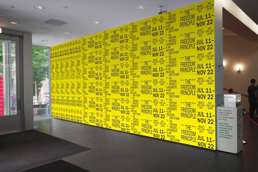 Bright yellow wall covered with rows of repeated black text stating the title and dates of the MCA exhibition *The Freedom Principle*