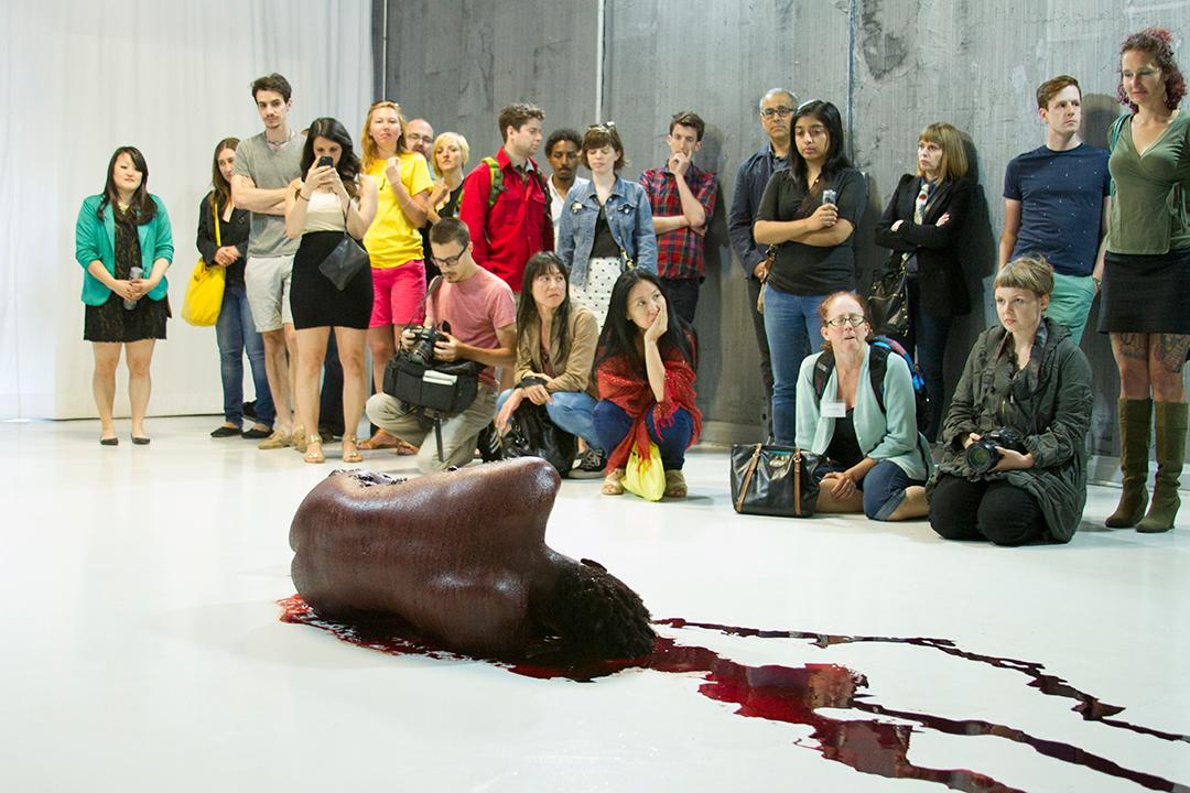 A naked performer smeared with blood is curled in the fetal position, facing a small audience.