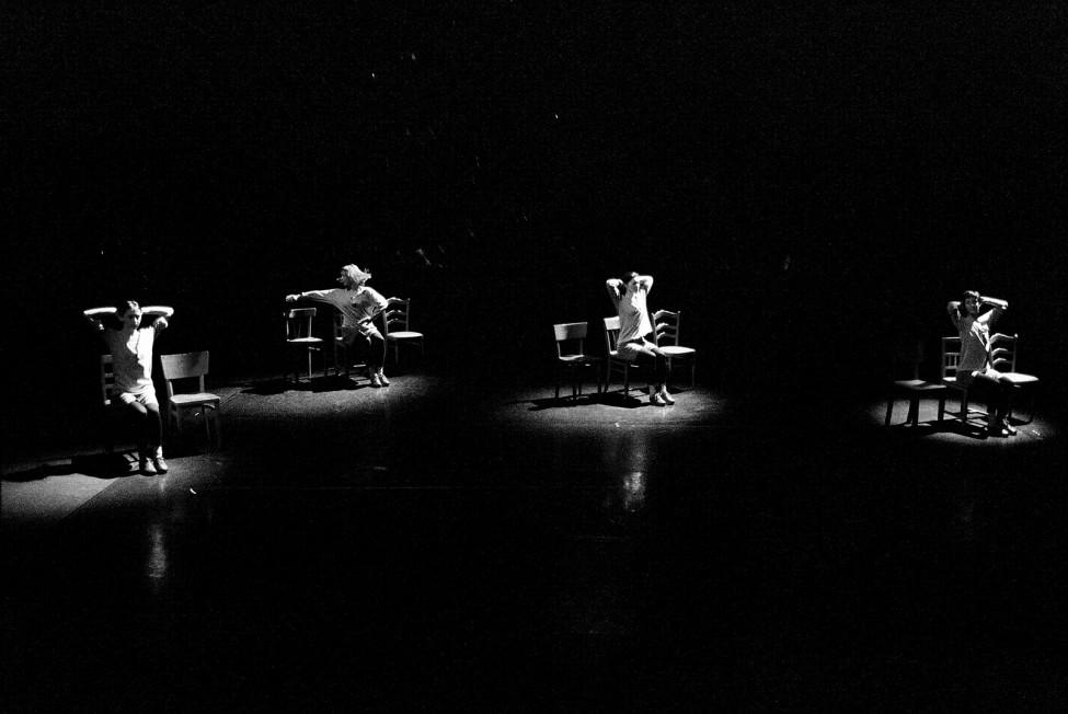 Black-and-white performance still of four women on a pitch-black stage, each one illuminated by a spotlight and dancing in a chair