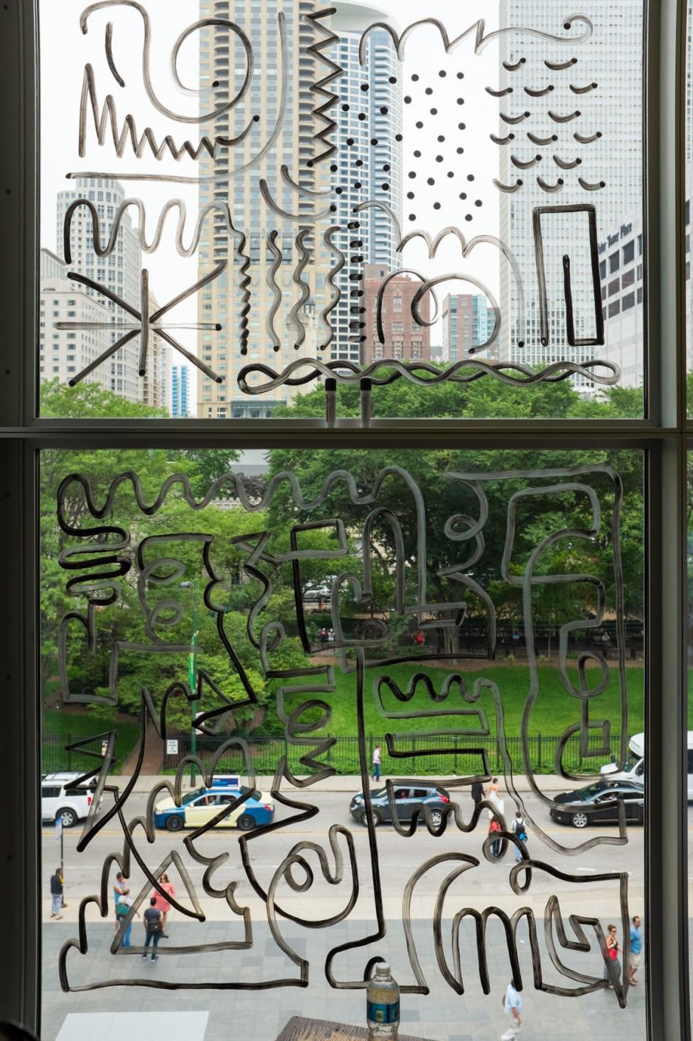 Various doodles in black marker appear on a window which looks out onto a lush park and tall buildings.