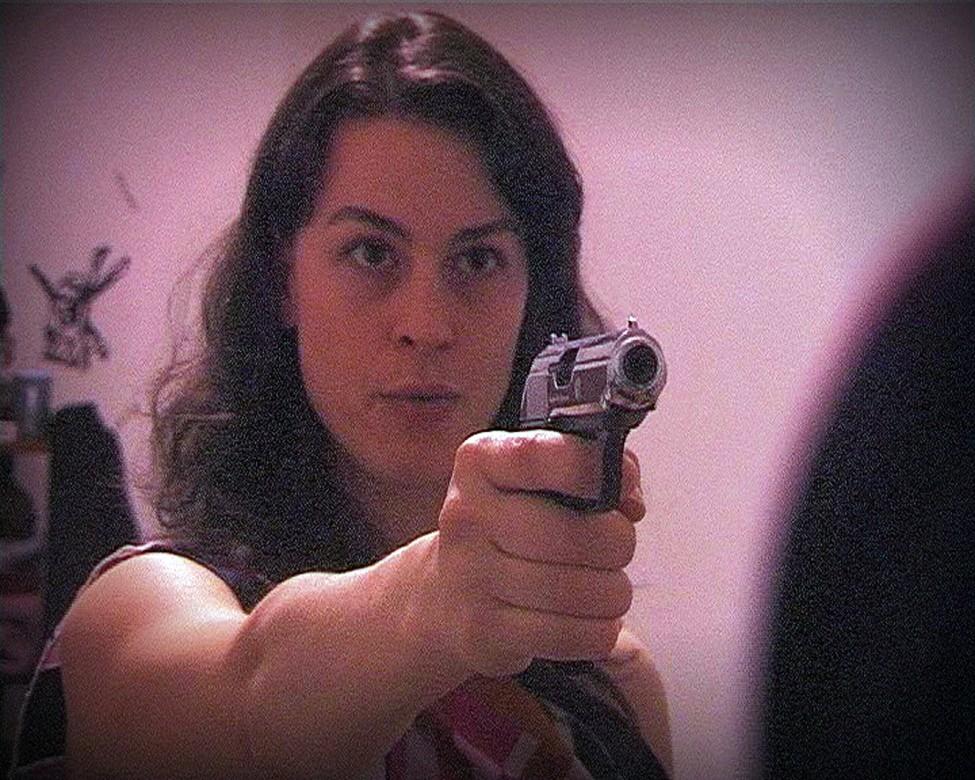 A magenta-tinged image of a woman with her arm outstretched aims a handgun just past you. Seemingly at someone whose presence is suggested by a dark silhouette to the right.