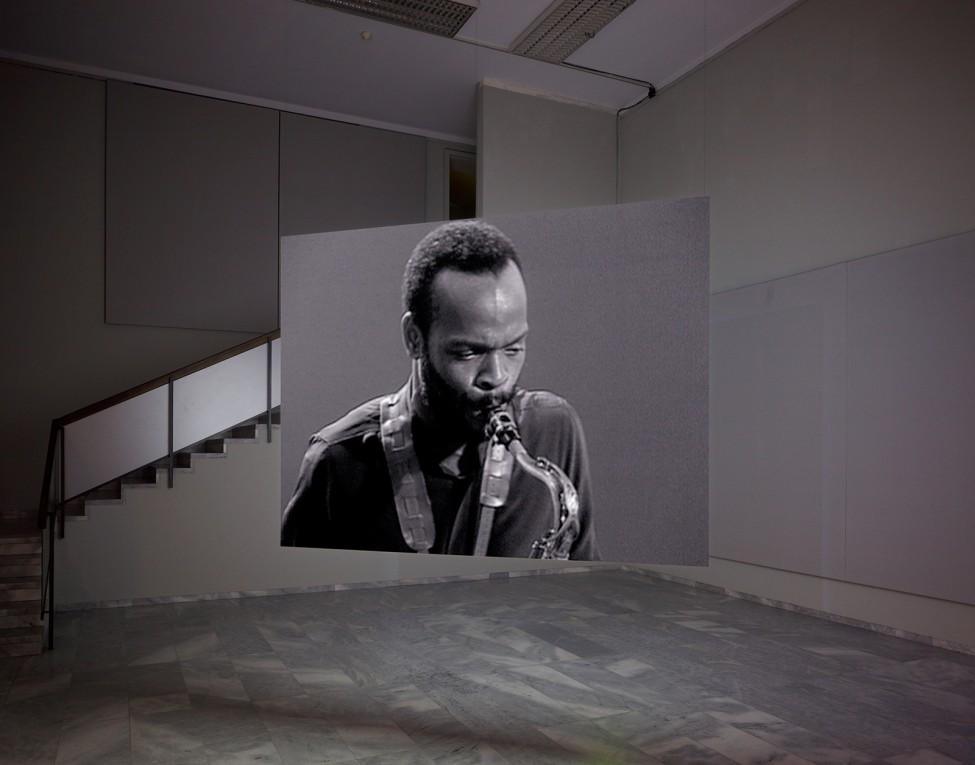 Installation view of a screen suspended from the ceiling with a video projection of a man playing the saxaphone