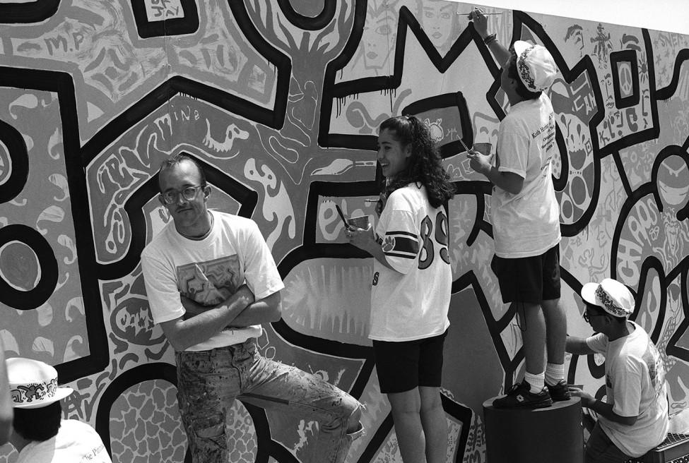 Teenagers in shorts paint a mural of graphic designs as a young man in glasses stands with arms folded looking at the camera.