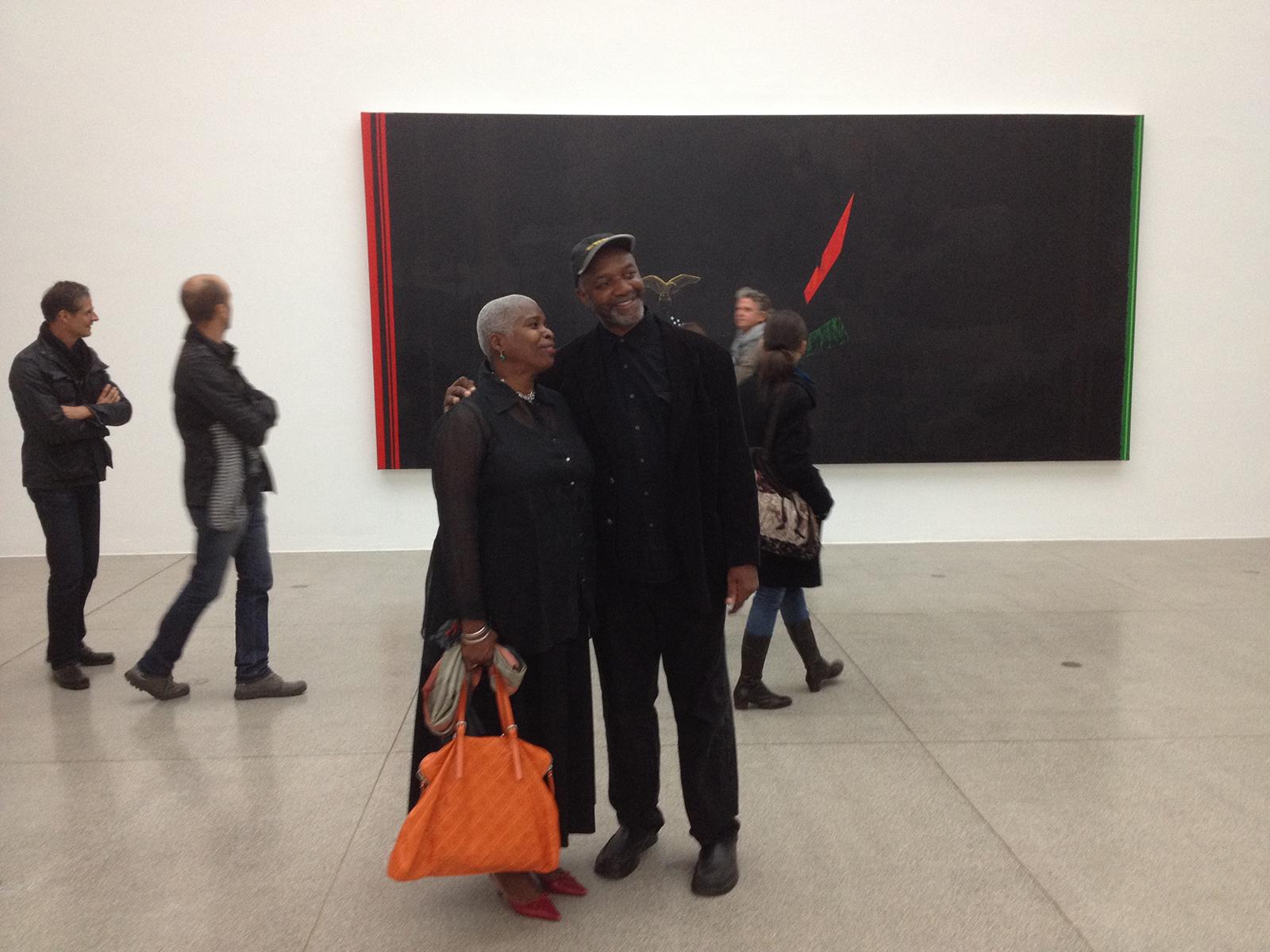 Two individuals smile arm in arm while standing in front of a large art piece.