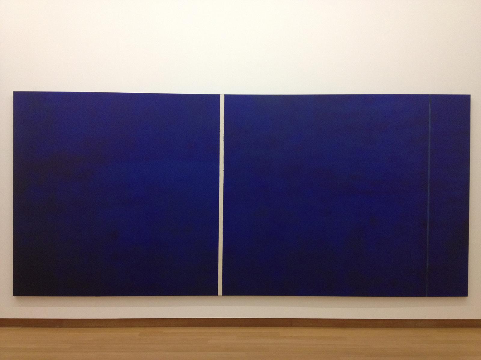 Installation view of a wide dark blue painting with a vertical white stripe running just left of center and a faint thin stripe near the right edge