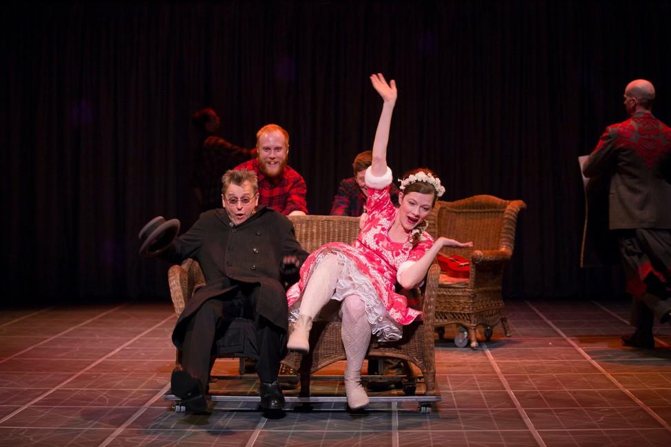 A man and a woman sit on a wicker bench with startled and amused expressions, as another man pushes the two individuals on the rolling bench along the stage excitedly.