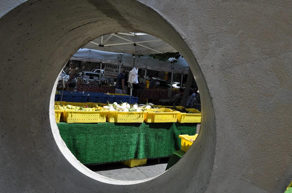 A table with yellow bins of vegetables seen through a circular hole in a large concrete sculpture