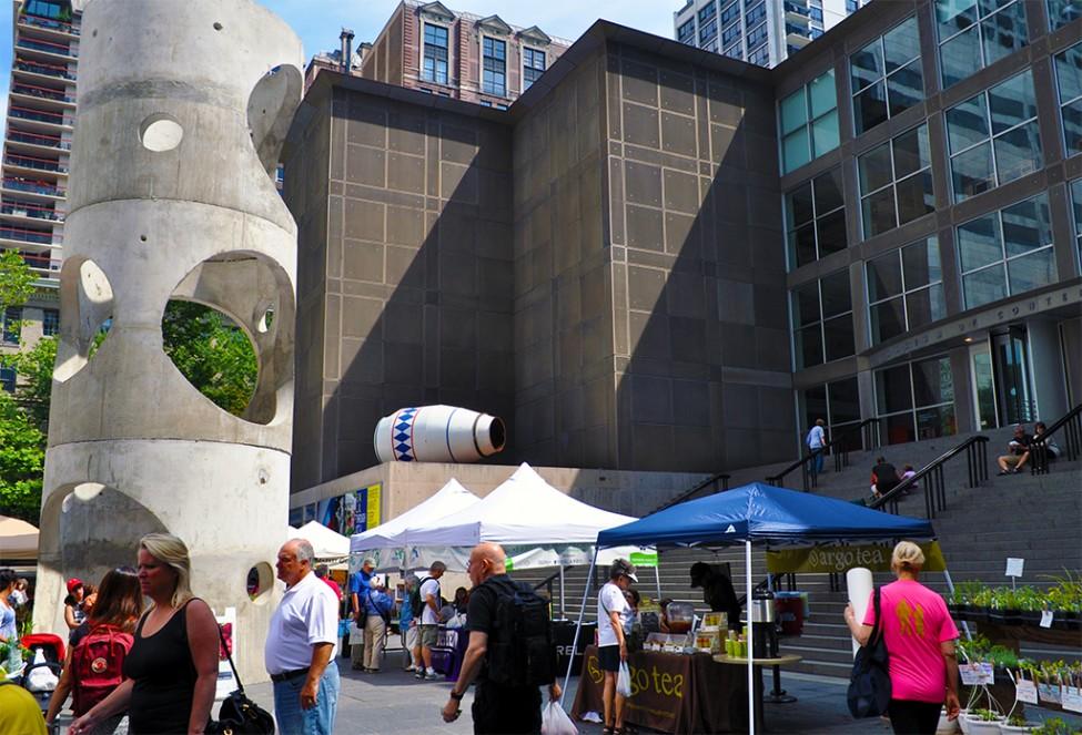 View of the MCA Plaza with two sculptures, a crowd of people, and farmers' market stalls