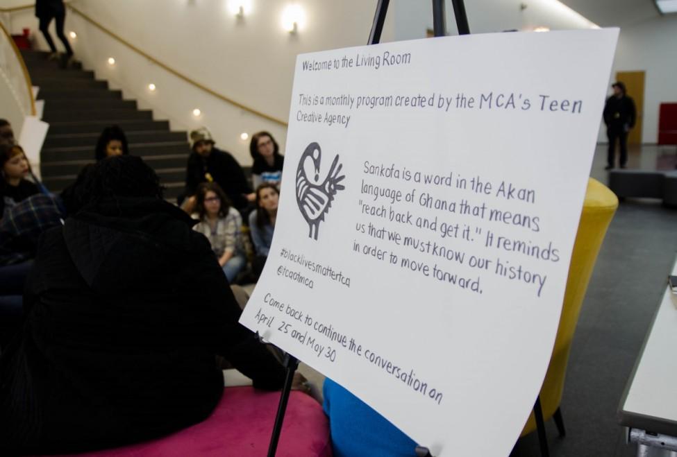 Out-of-focus teens sit in a circle in the MCA's education lobby behind a sign with the headline "Welcome to the Living Room."