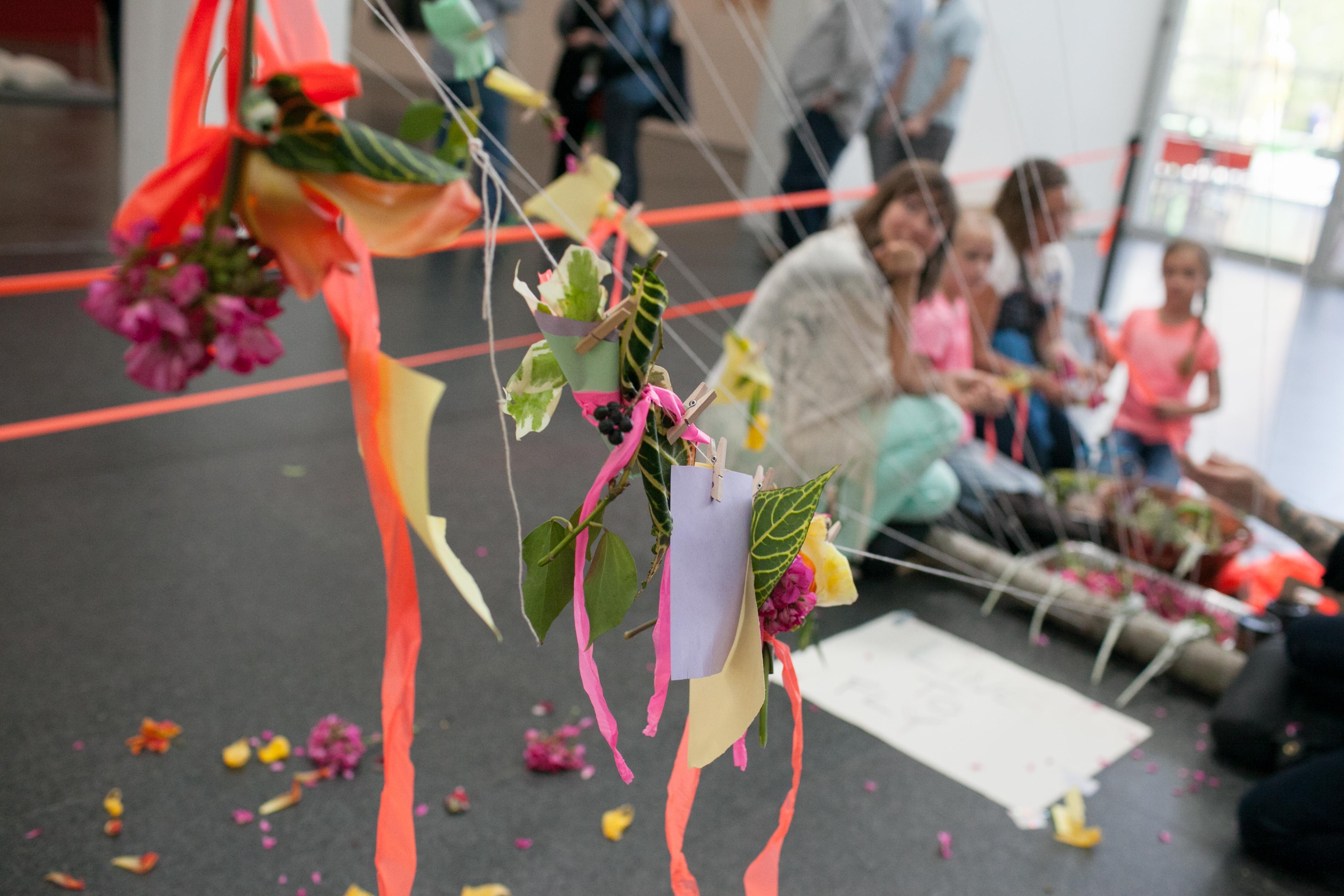 Adults and kids look through an installation of fake flowers and ribbons attached to strings with clothespins in the MCA's Atrium.