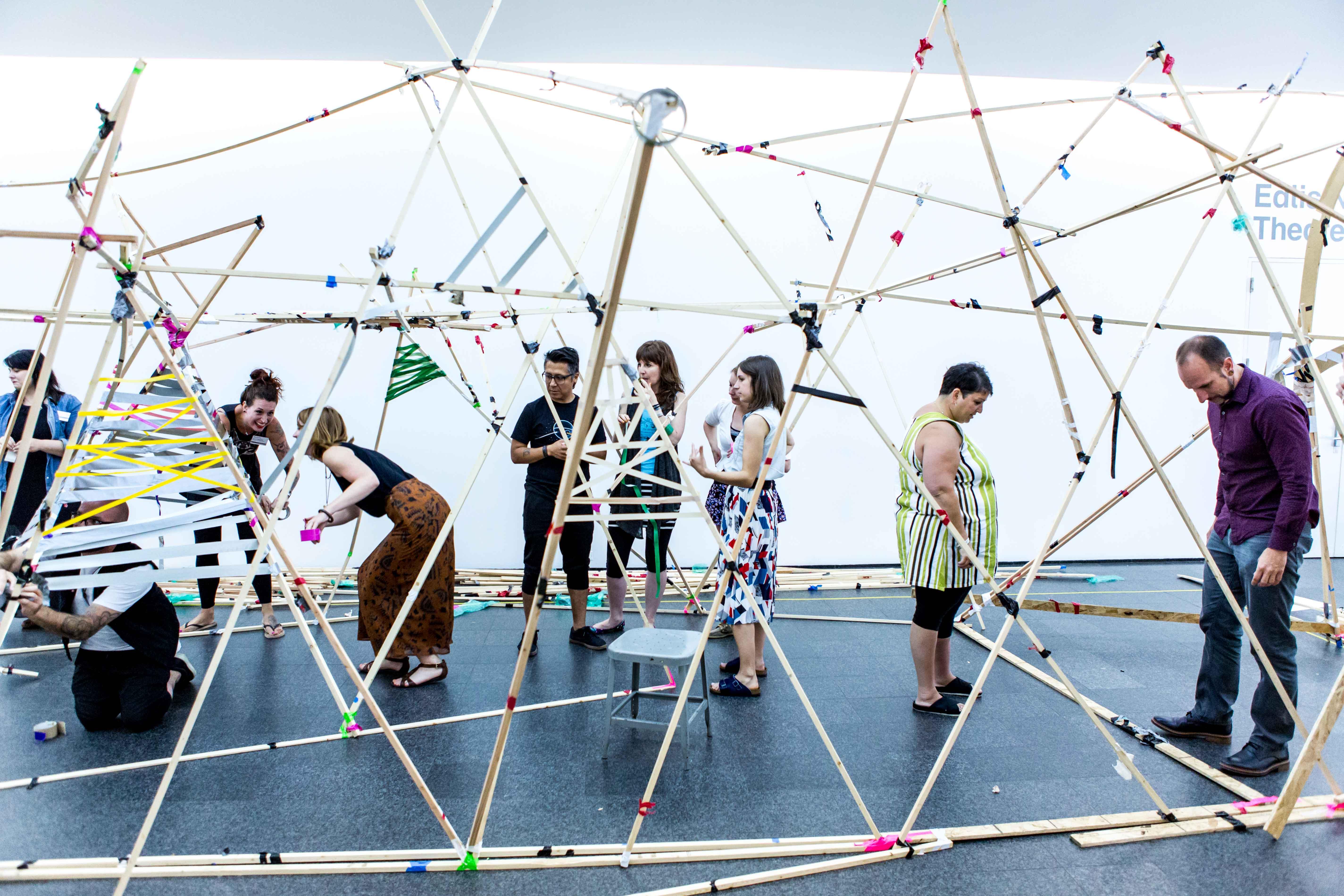 Adults standing in an open-air structure made with slender wooden beams and colorful tape