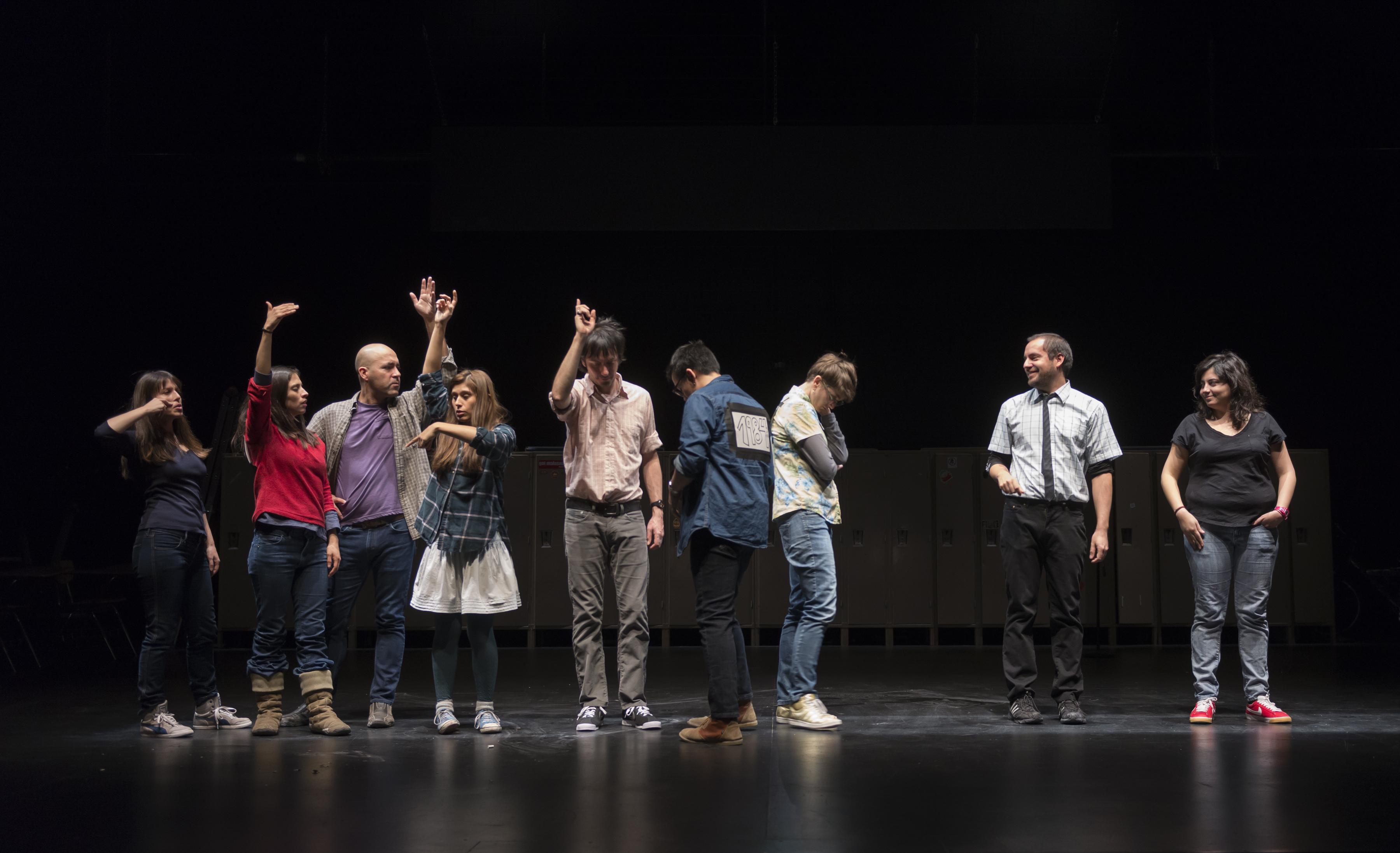 On a dark stage with lockers barely visible in the background, nine people stand in a row, five of them gesturing with their arms.