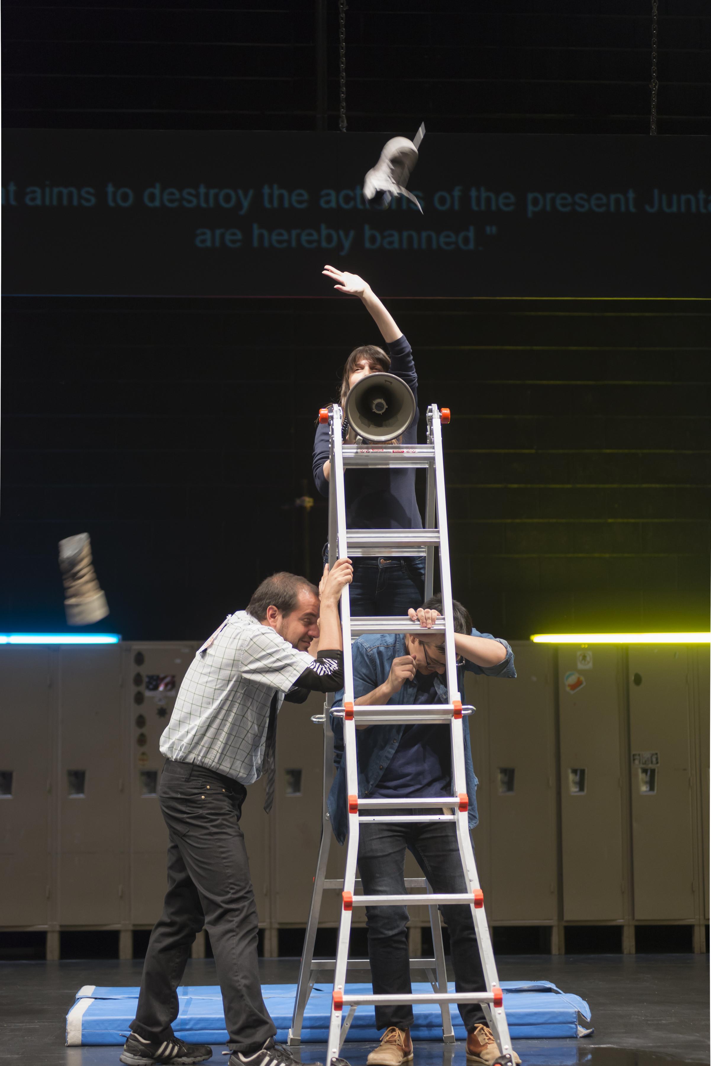 On a stage set with lockers and gym mats, a woman on a freestanding ladder talks through a loud speaker. She has thrown something in the air, and two men below her cringe, taking shelter under the ladder.