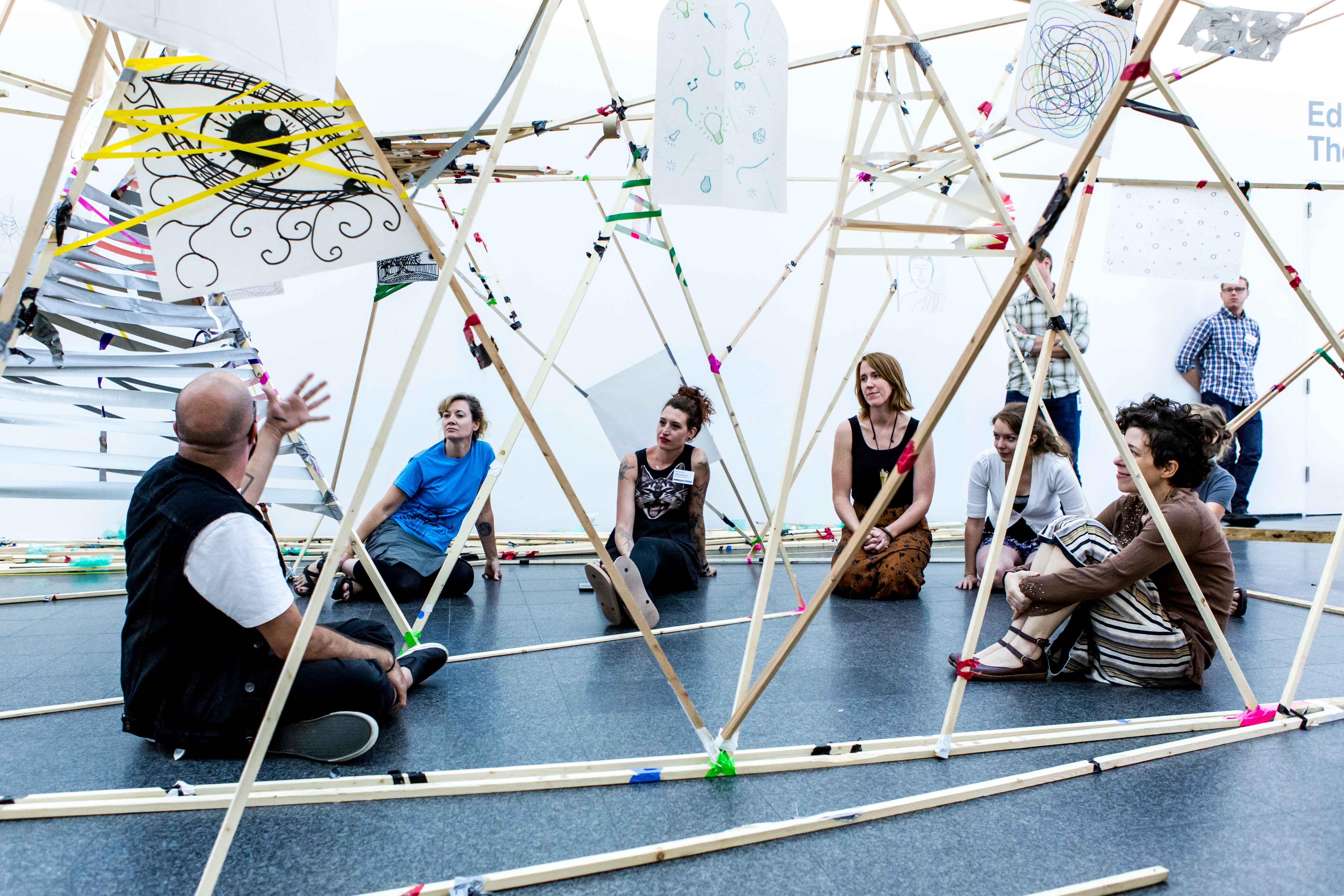 People sit on the ground in an open-air structure made of long wooden beams and tape