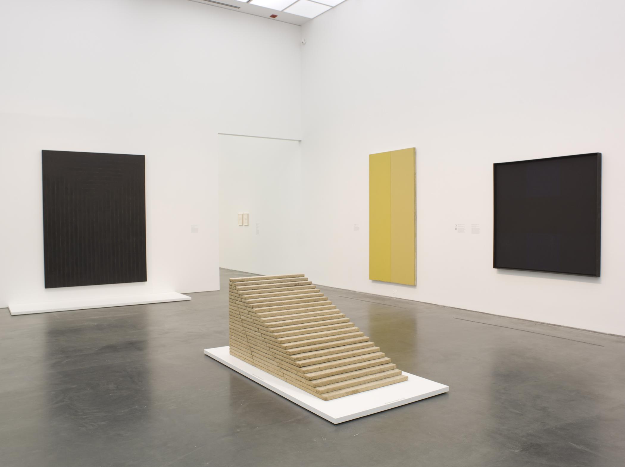 A gallery view with minimalist color field paintings on the walls, and a stairway-like sculpture in the middle of the room.