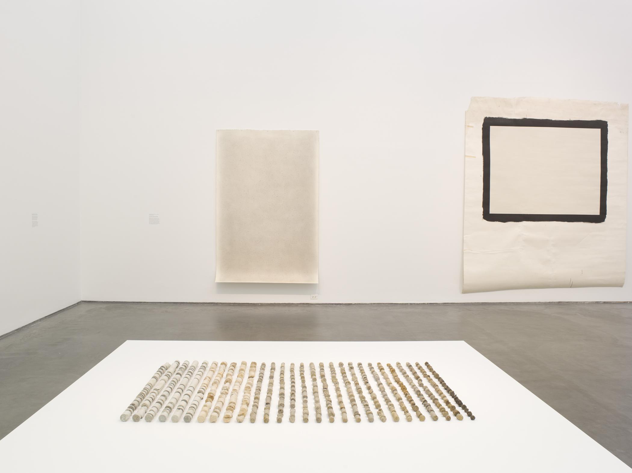 Installation view of two paintings and a sculpture composed of rock fragments arranged in rows and organized by color on a white platform