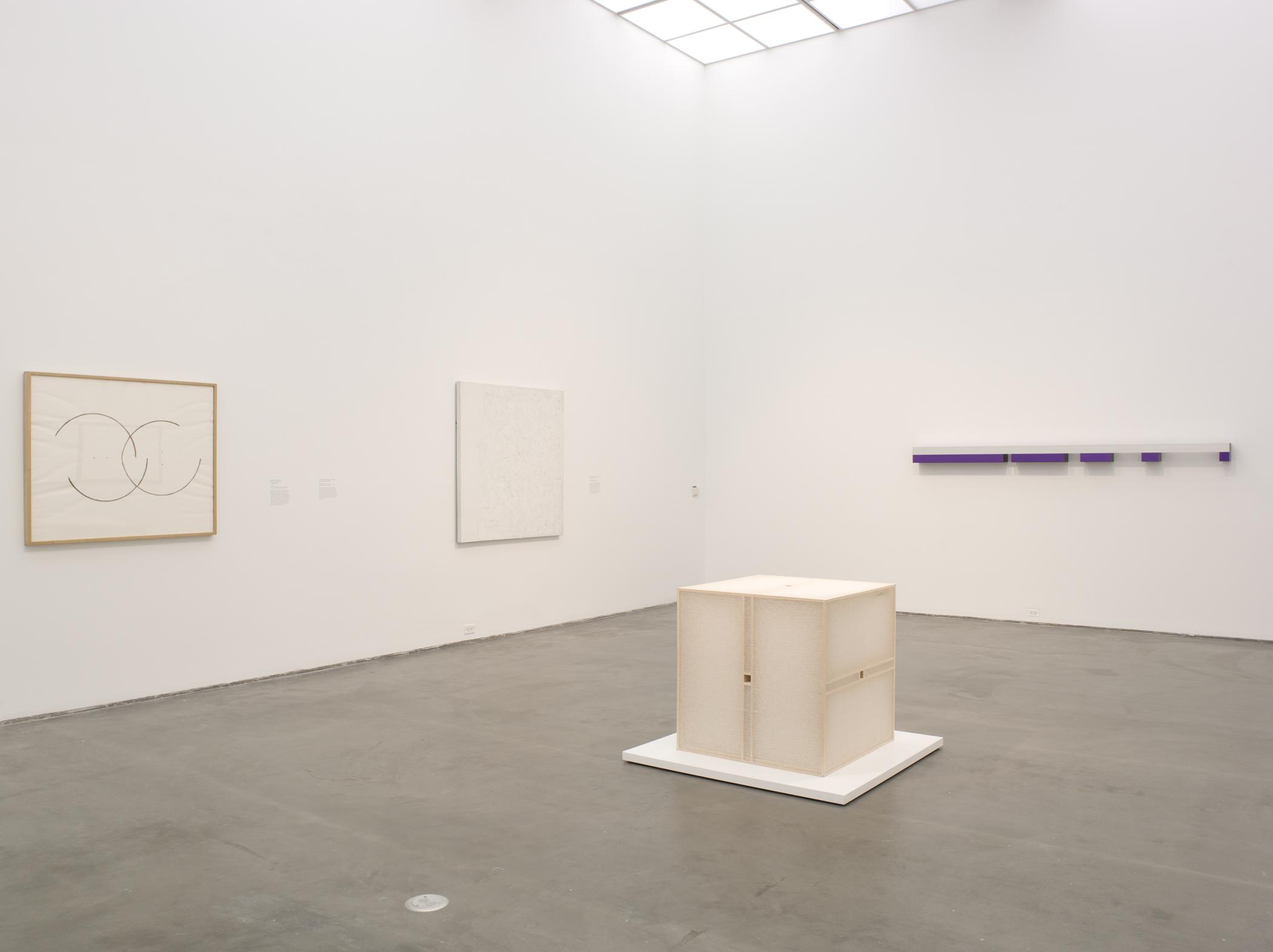 Installation view of a cube-shaped sculpture on a white platform, two paintings, and a wall sculpture