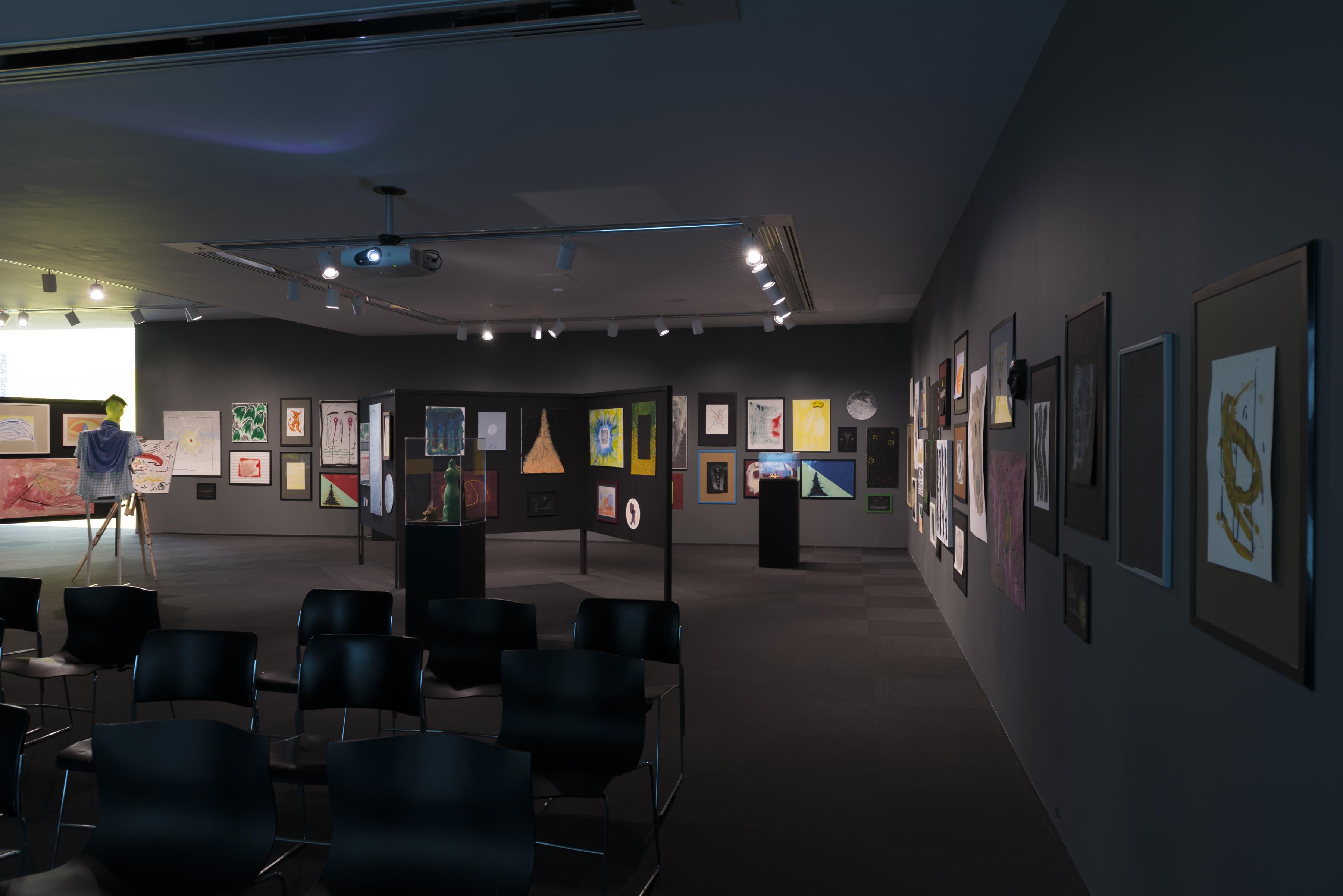 Installation view of paintings and sculptures in a grey gallery, rows of chairs in the foreground, and a humanoid figure on the left standing at an easel and canvas