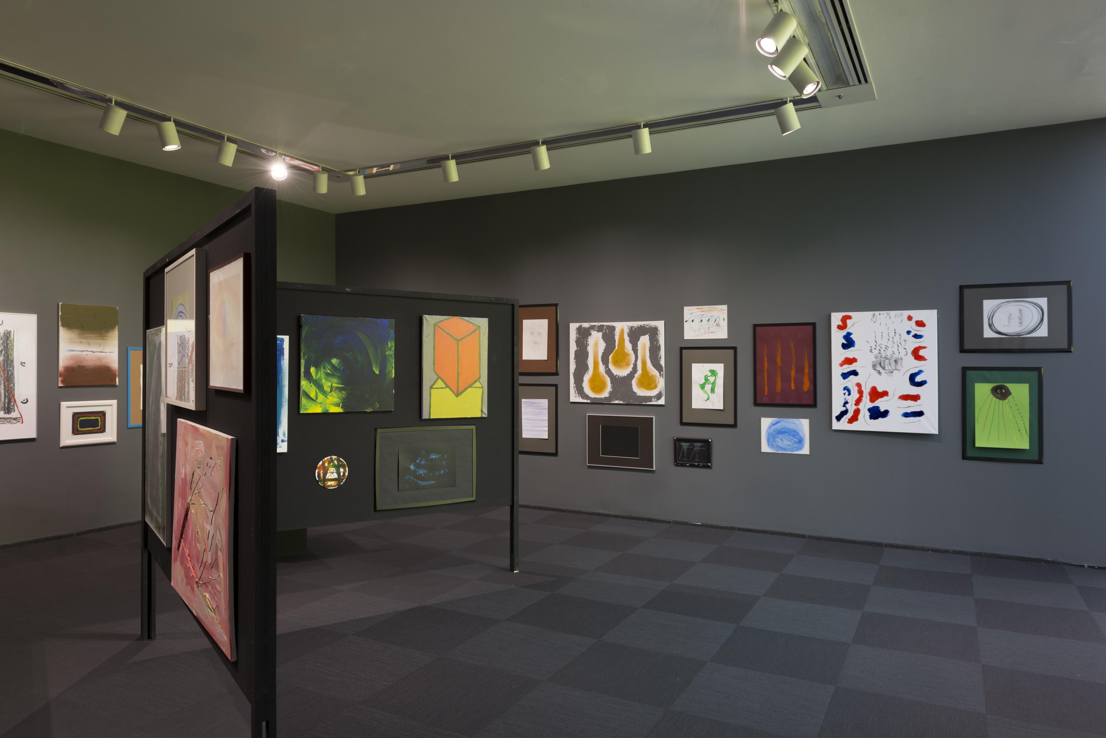More than a dozen paintings and three sculptures on a pedestal in a grey gallery with a black folding display board