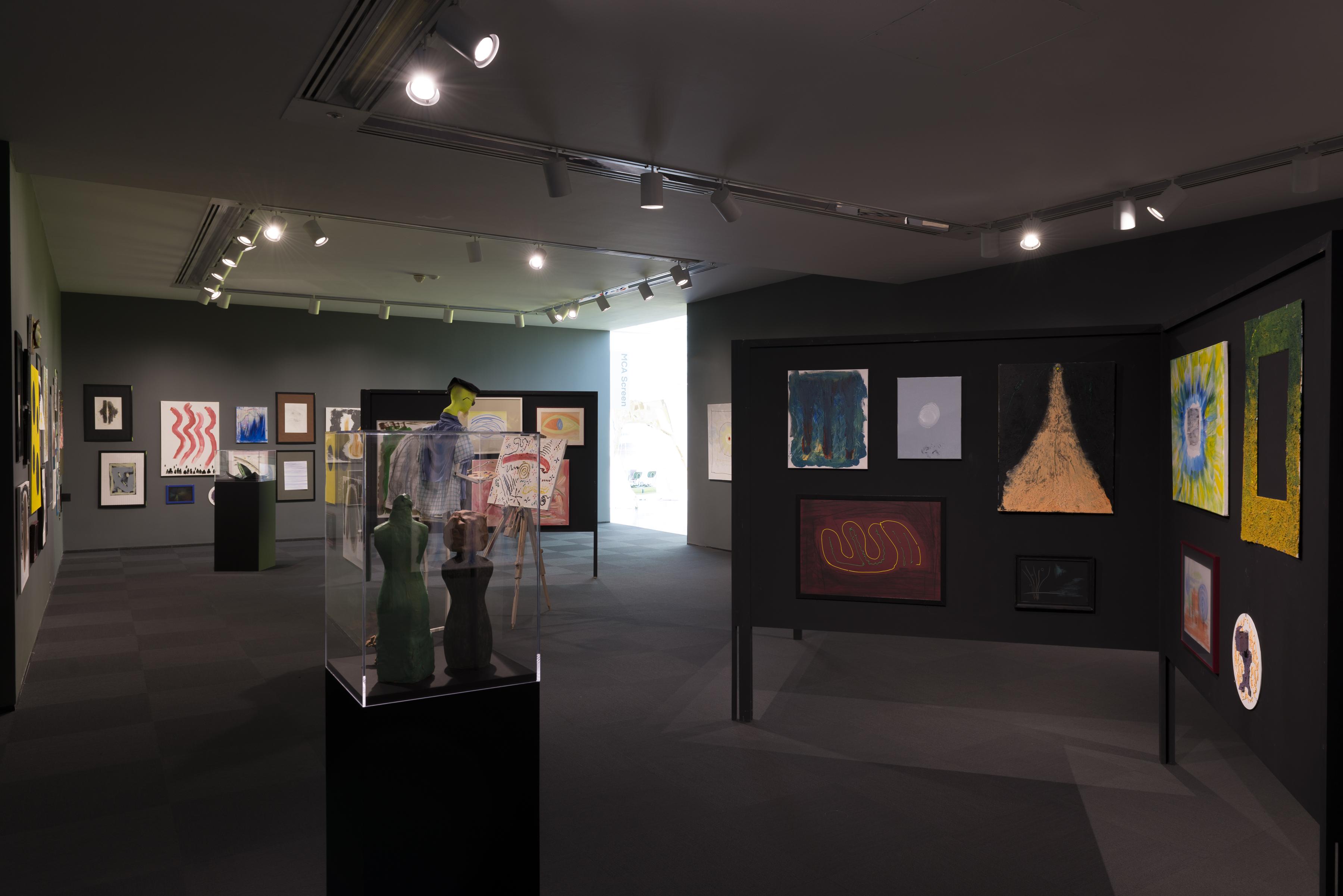 Installation view of sculptures on pedestals, colorful abstract paintings hung in clusters on a folding display board and on the walls, and a humanoid figure painting at an easel in the middle of the room