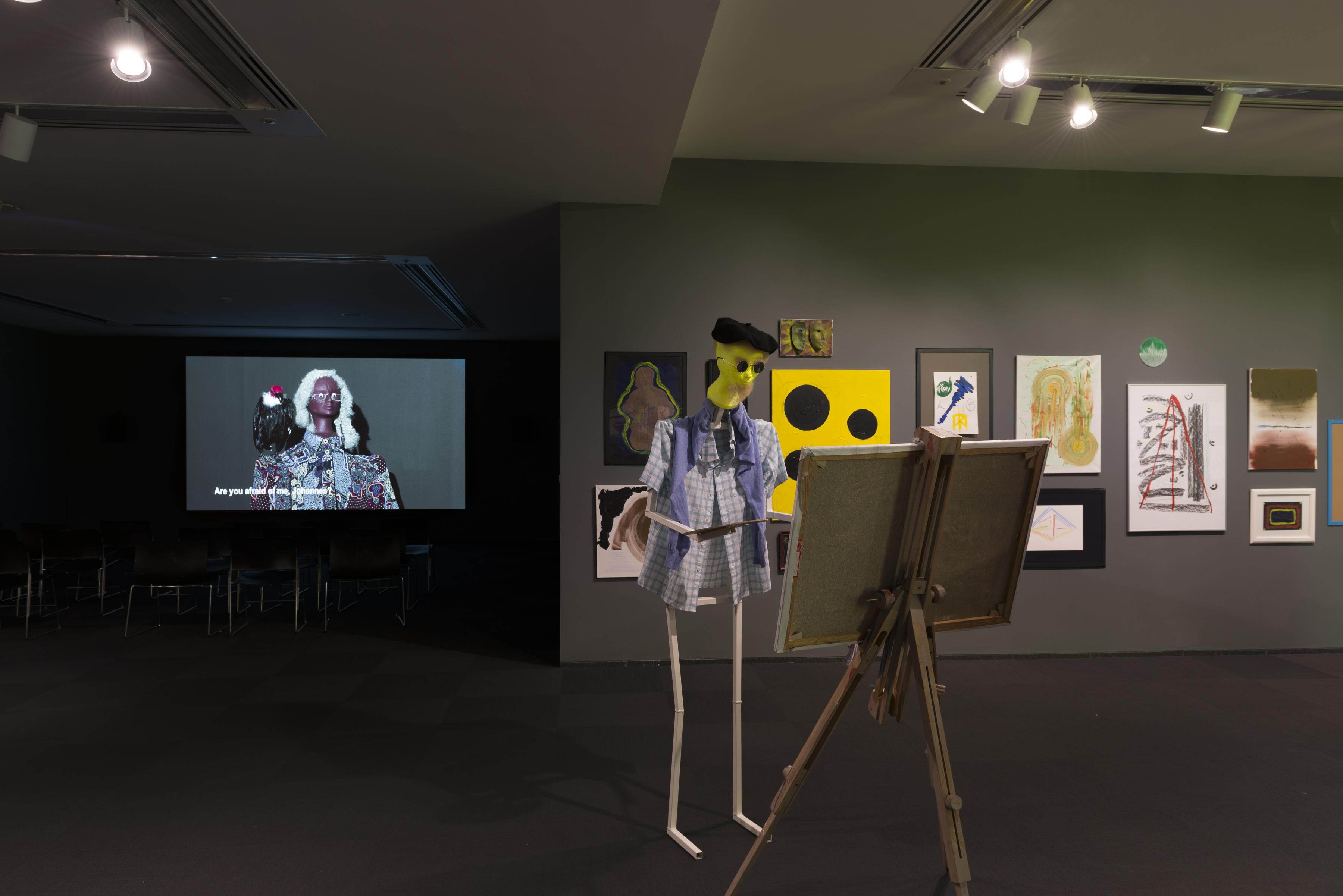 A humanoid figure wearing a beret and plaid shirt stands at an easel holding a paint palette. There are abstract paintings and a video projection of a human doll in the background.