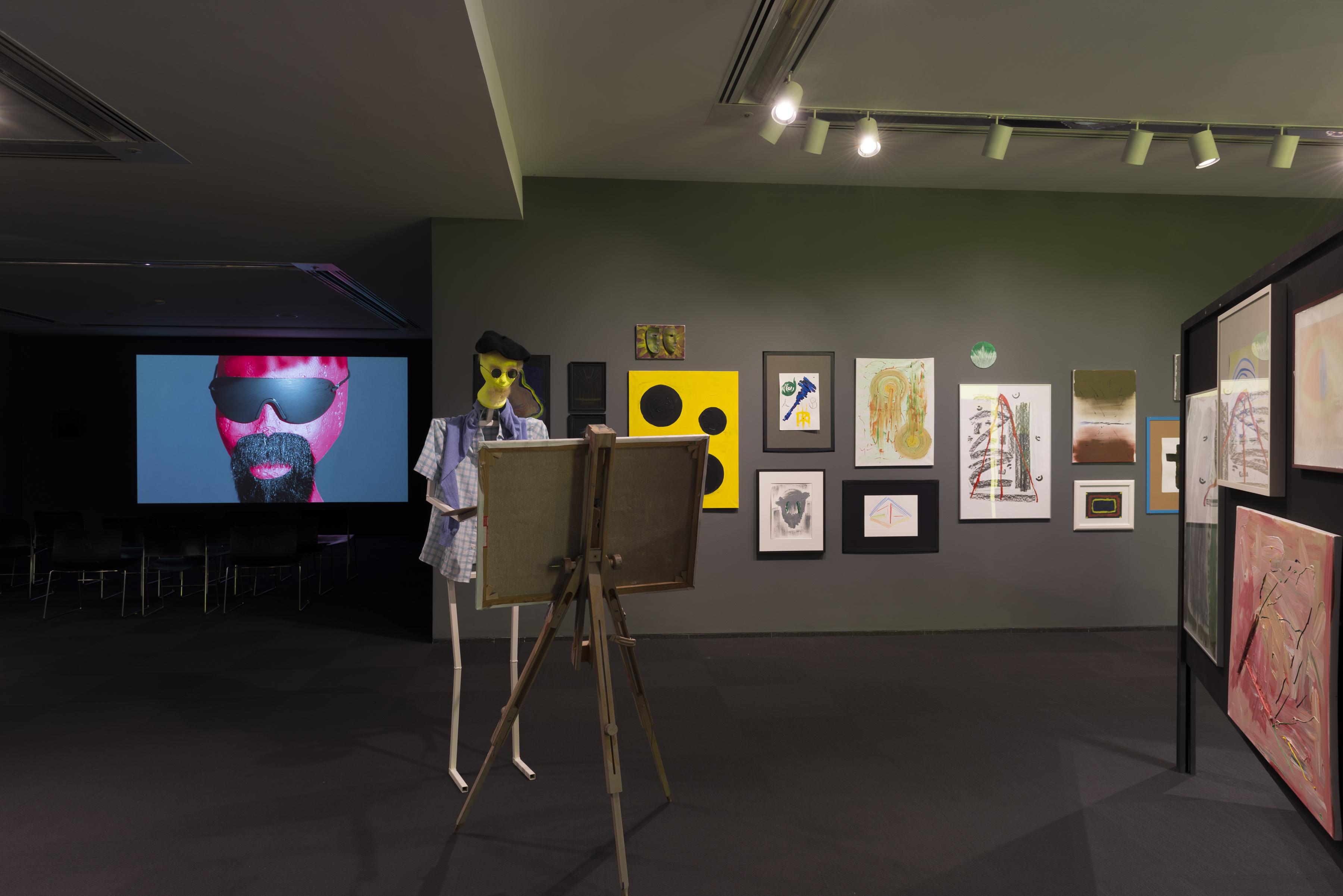 A humanoid figure wearing a beret and plaid shirt stands at an easel holding a paint palette. In the background, there are abstract paintings hung in a cluster on the wall and a video projection of a human doll with a goatee and wearing sunglasses .