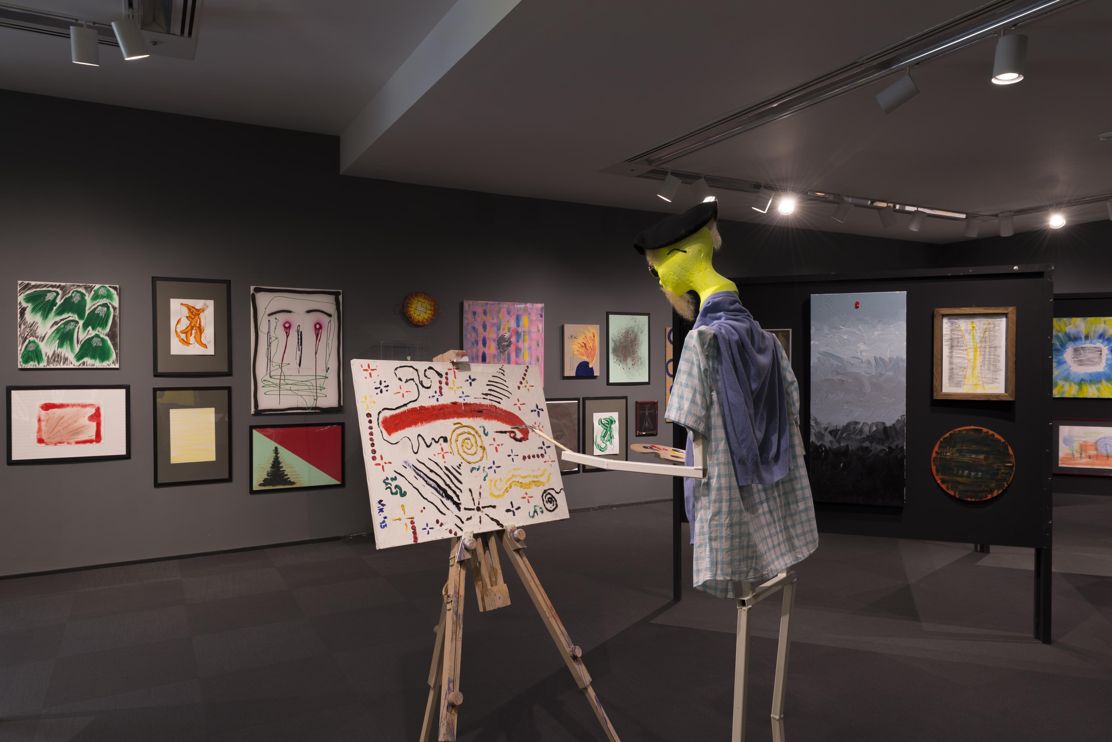 In this photograph, a humanoid sculpture with a yellow head wears a plaid shirt and paints a canvas on an easel in the middle of gallery densely hung with paintings.