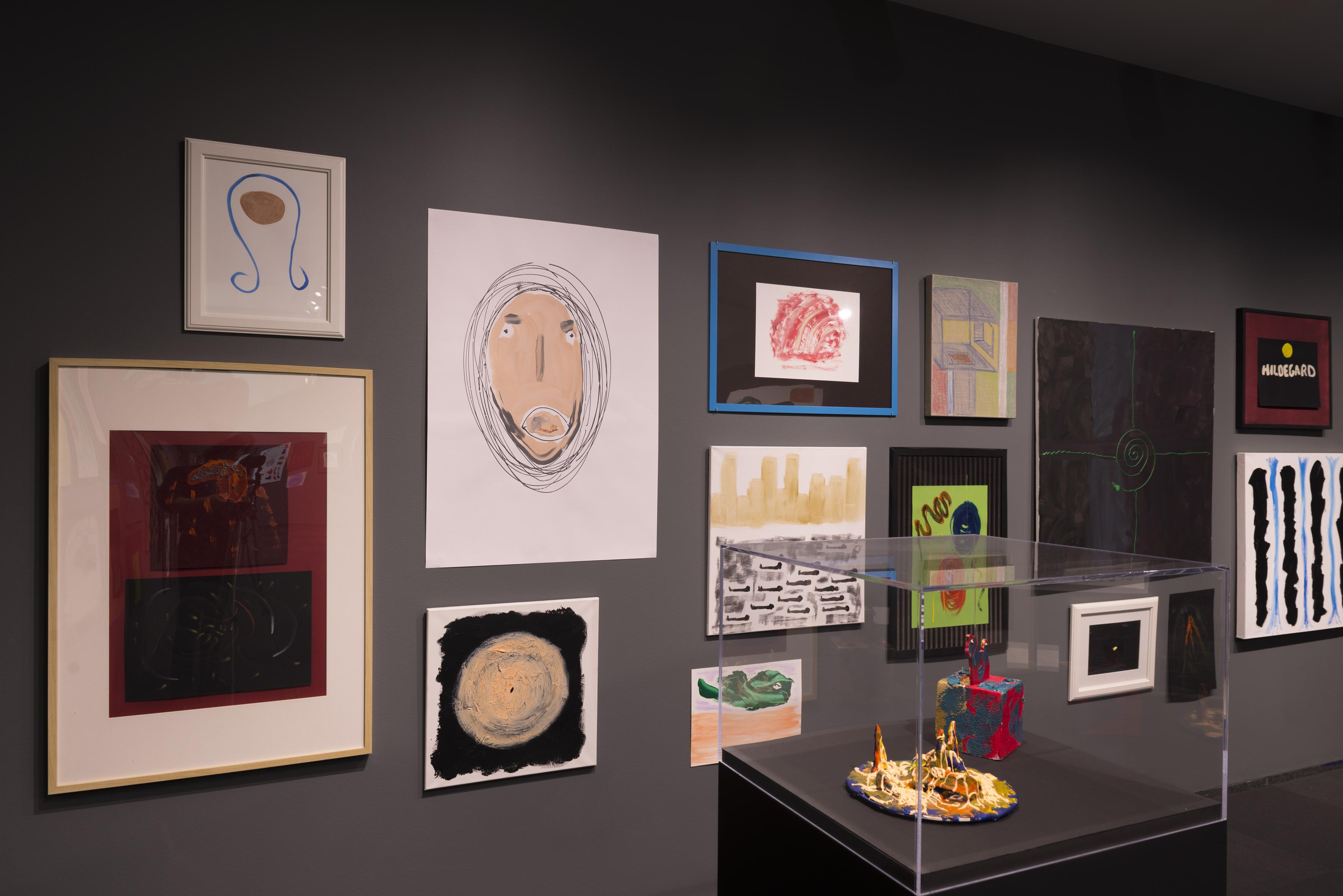 Installation view of a cluster of colorful drawings and paintings hung on a grey wall and a pedestal with two sculptures in a clear, protective case