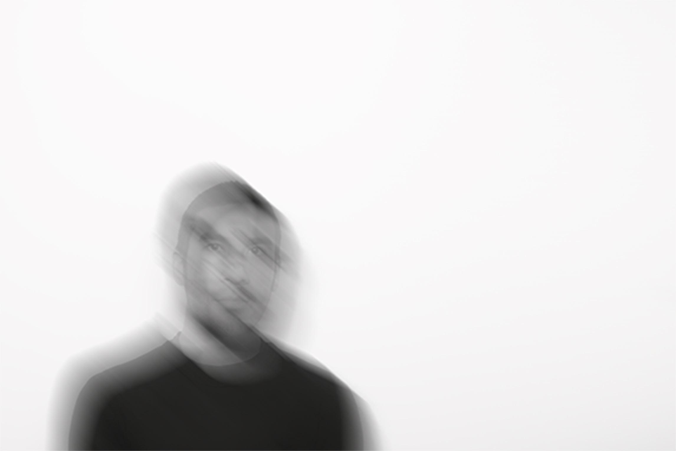 A blurry, black-and-white portrait shows a young man with buzzed dark hair against a white background.