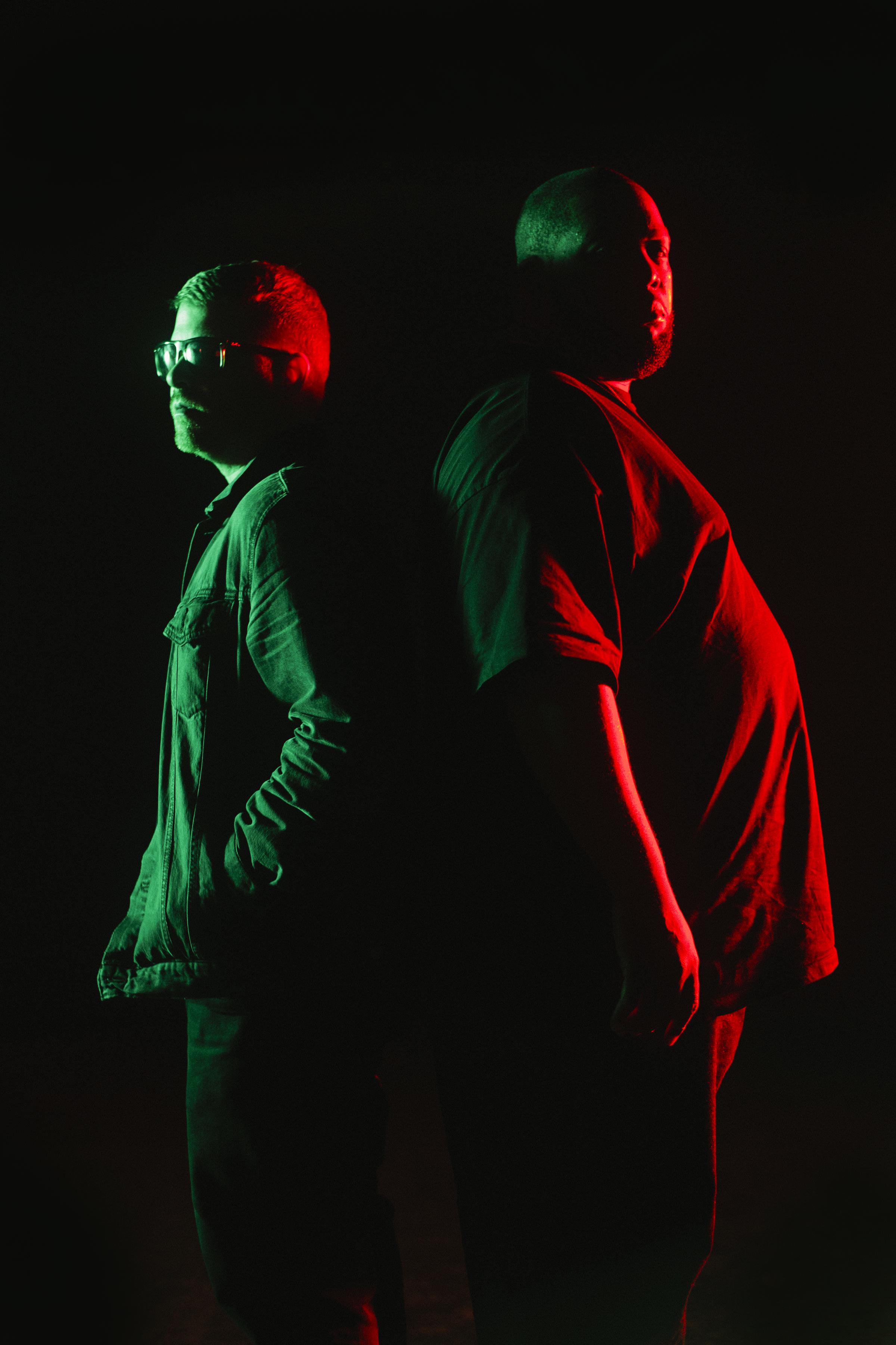 Two men standing back to back in a pitch black room are partly illuminated by a green and red light.